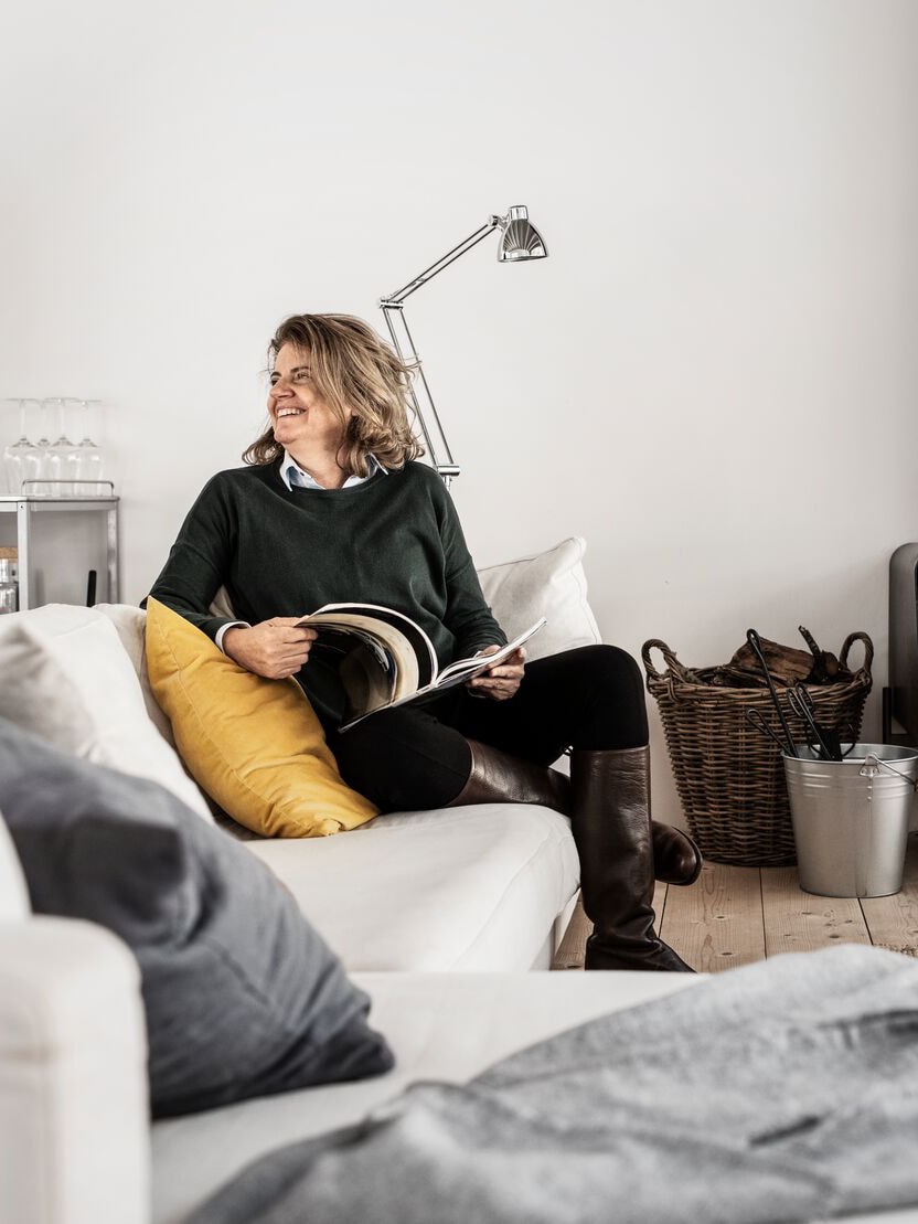 A woman with a smile on her face is sitting in a sofa in the living room while reading a magazine.