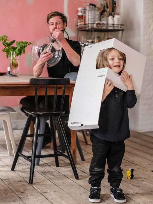 A father is sitting by a dining table assembling a lamp while his son is playing with the cardboard box.
