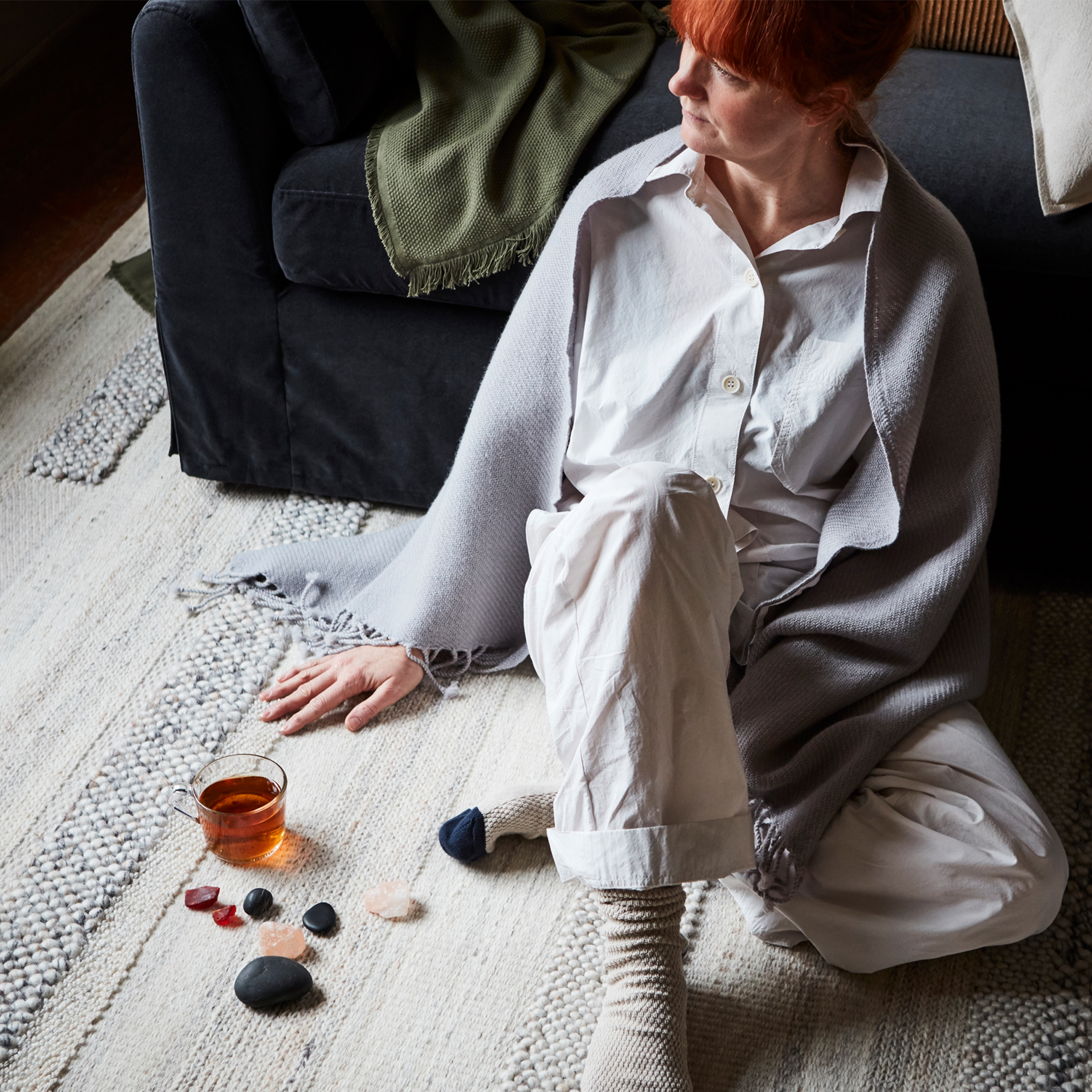 一位红色头发的女士倚靠着沙发坐在地毯上,肩上披着一条披肩,身旁放着