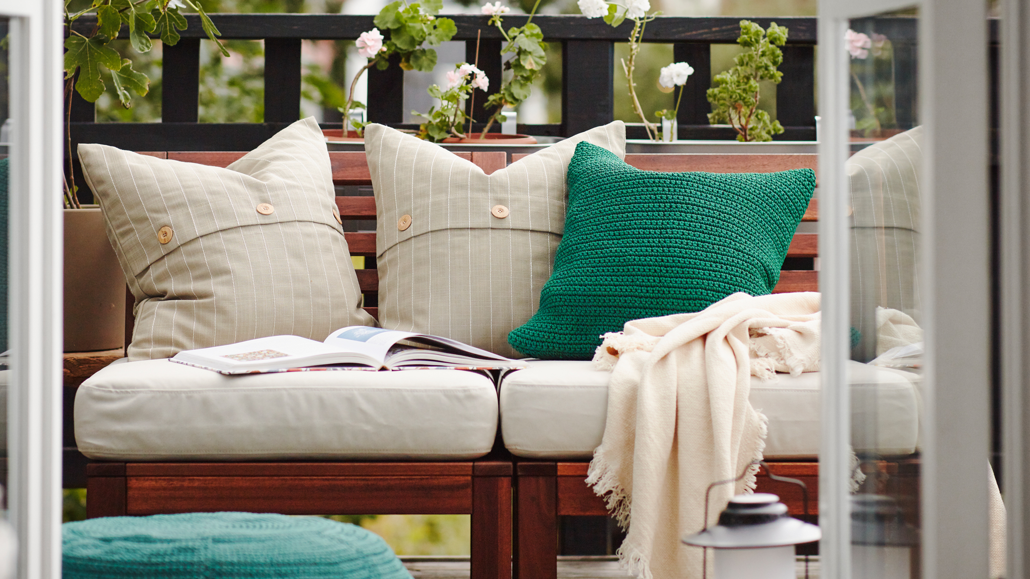 A balcony with a wooden outdoor sofa with white and green cushions, a throw and an open book on it, green plants behind.