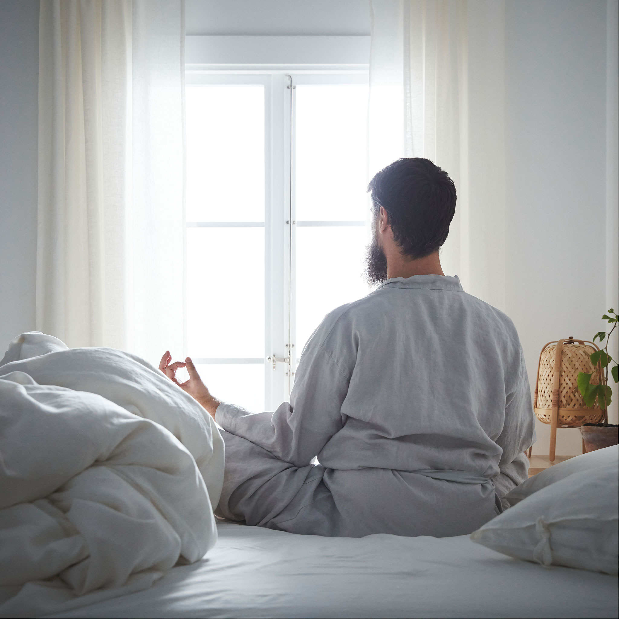 A bearded man in a calm-looking room sits legs-crossed in a meditation pose, his back to the camera.