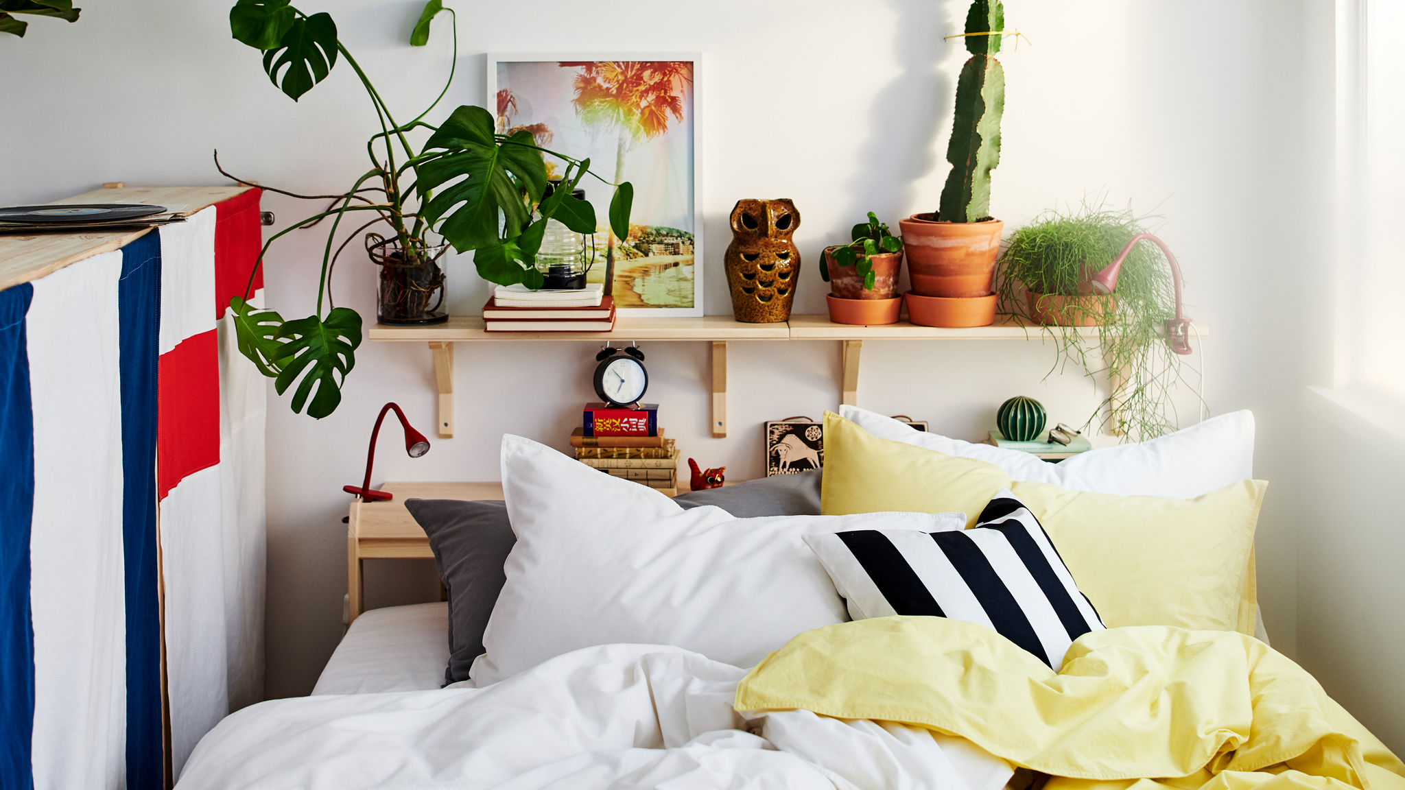 A bed between a window and the back of a storage unit with lots of pillows and yellow, white, black and grey bed linen.