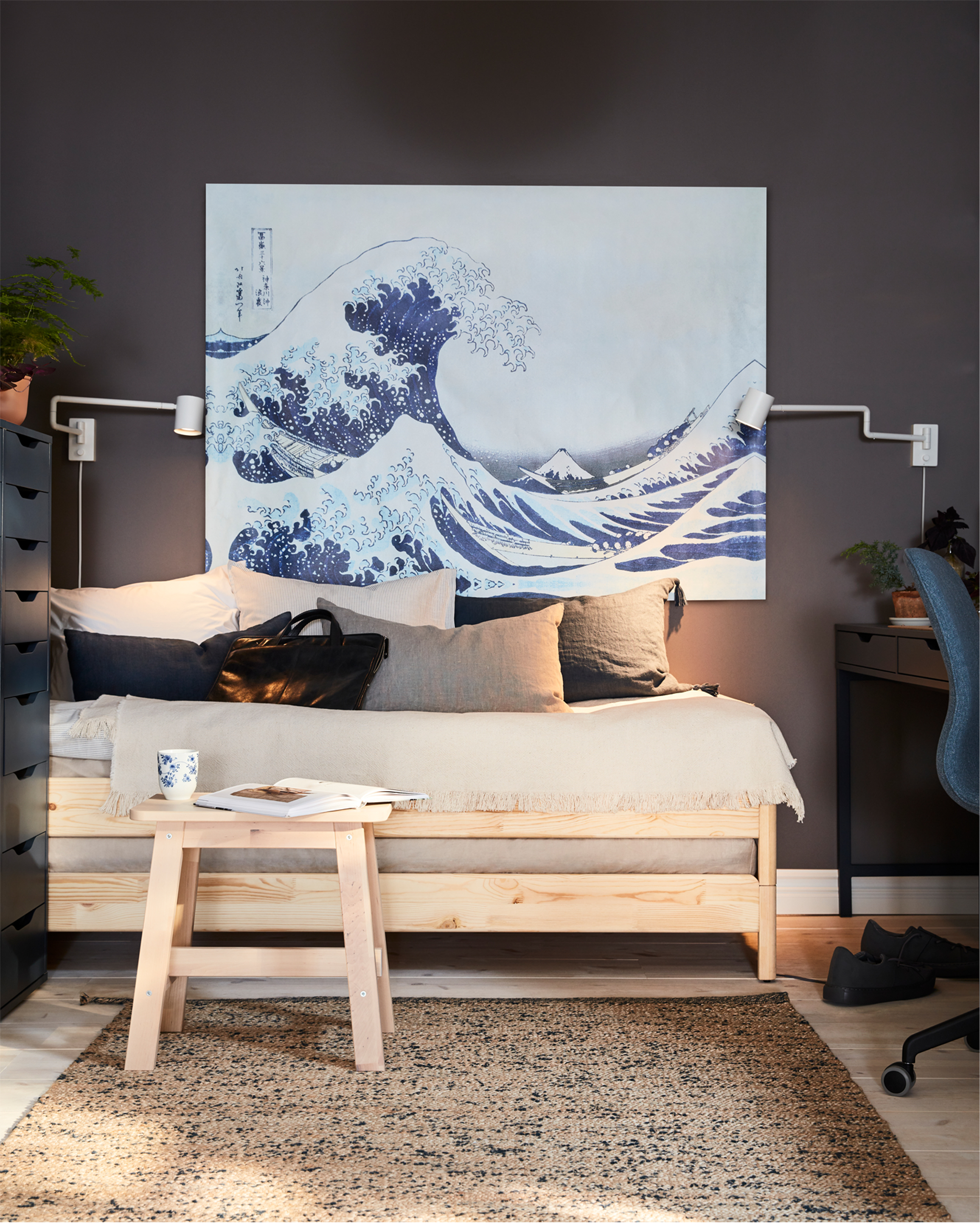 A bed in pine and a birch stool with a book and a mug in front, a big painting with a wave and two white wall-mounted lamps.
