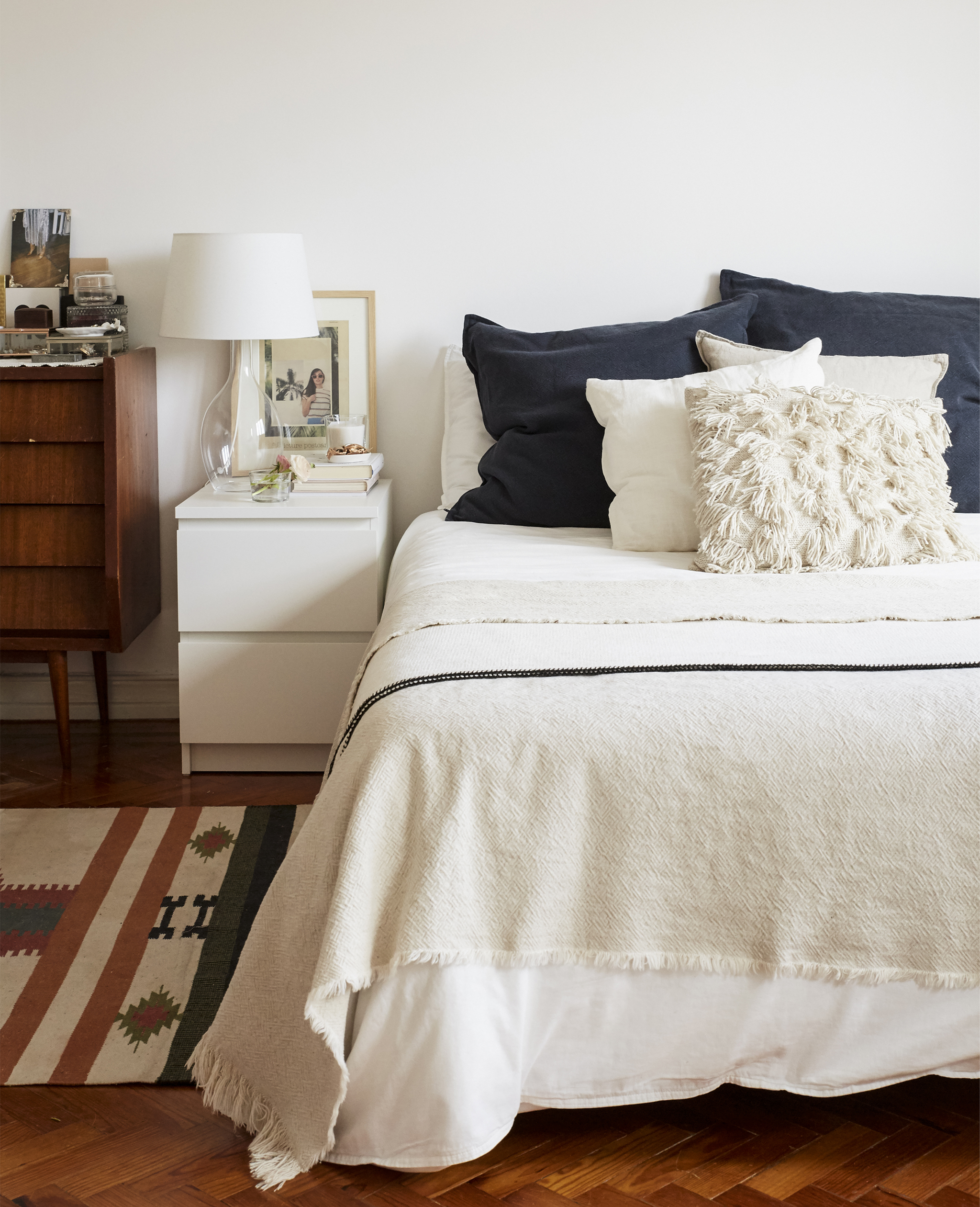 A bed with white bedding and navy cushions, a modern bedside table and a vintage cabinet.