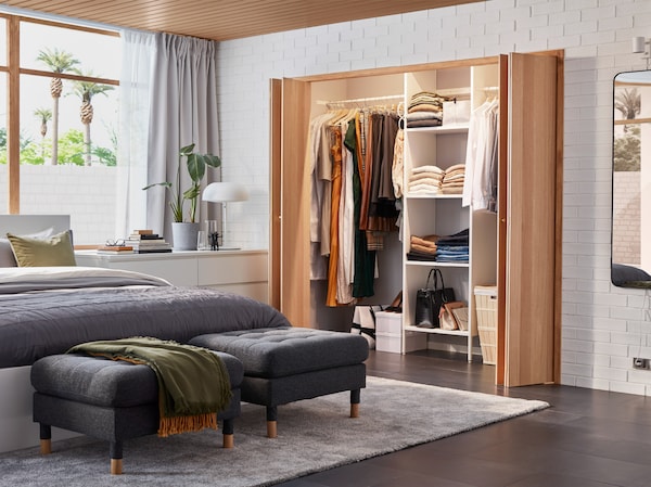 A bedroom with a built-in wardrobe solution, a white bed frame, a white chest of drawers and two dark grey footstools.