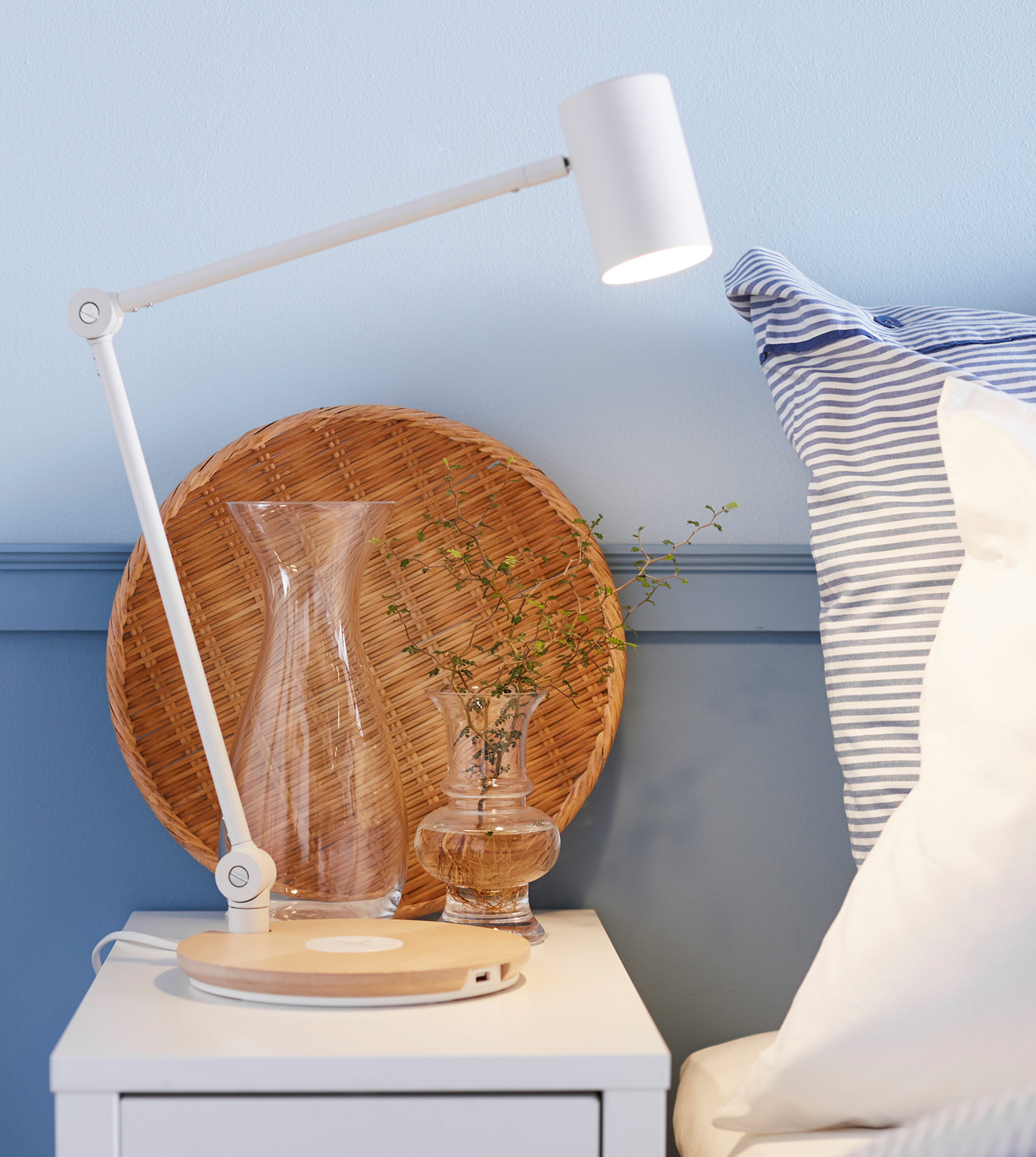 A bedside table is set with a lamp, vase of small flowers and a wicker tray.