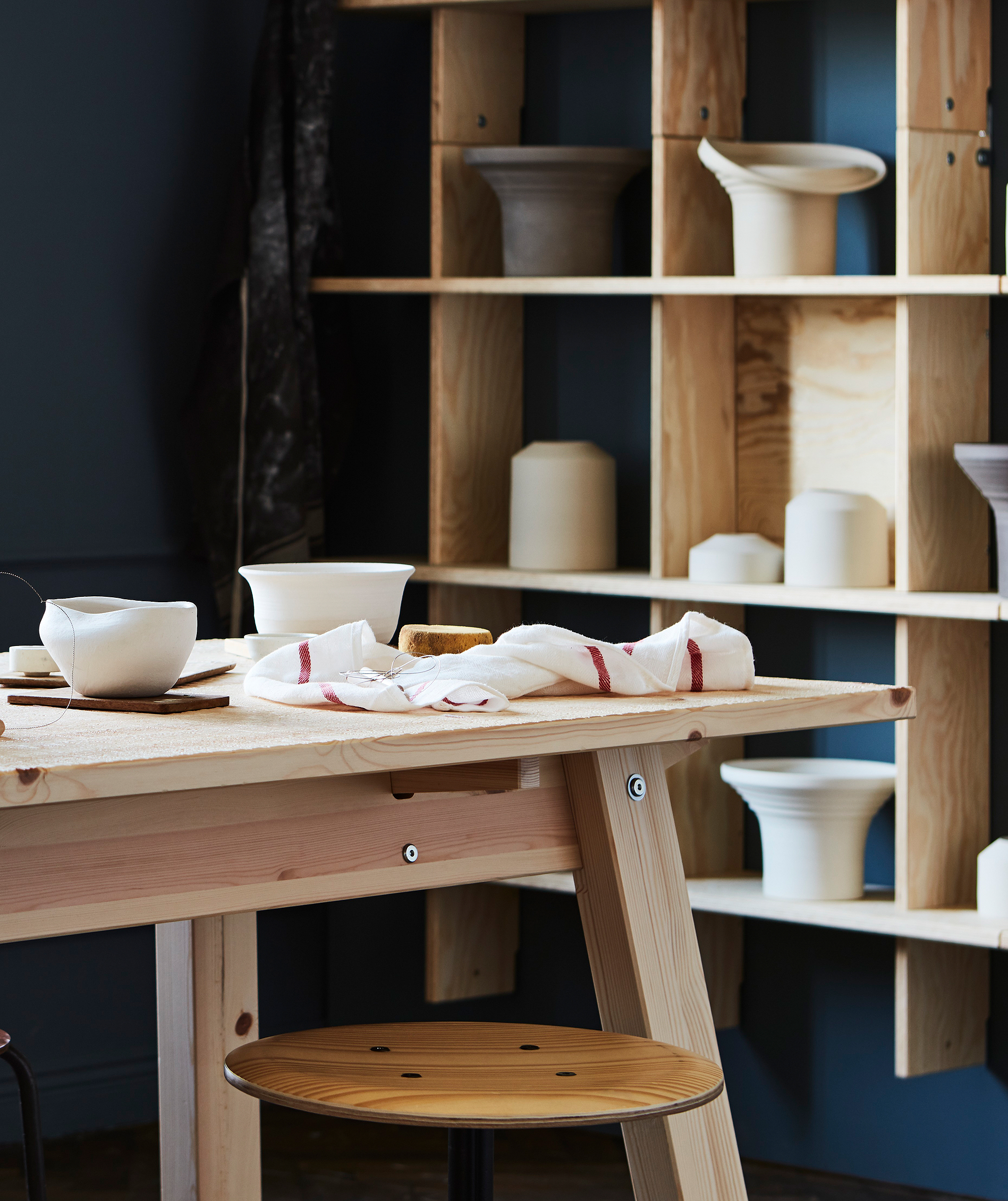 A big table in untreated, blond wood in a room painted in rich colours; large blond-wood wall shelf with pottery in back.