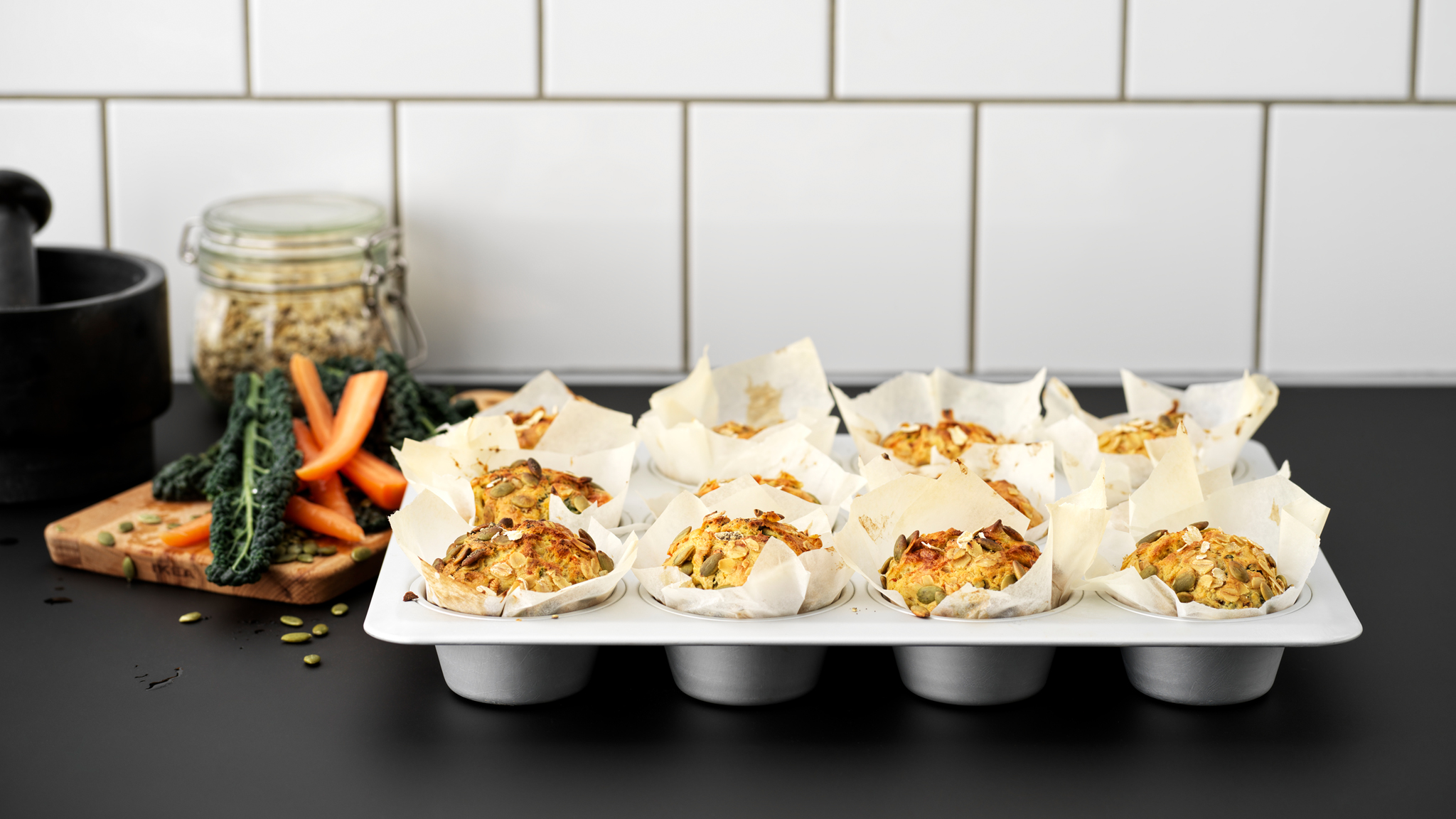 A black kitchen worktop with a VARDAGEN metal muffin tin filled with twelve muffins made with carrots and pumpkin seeds.