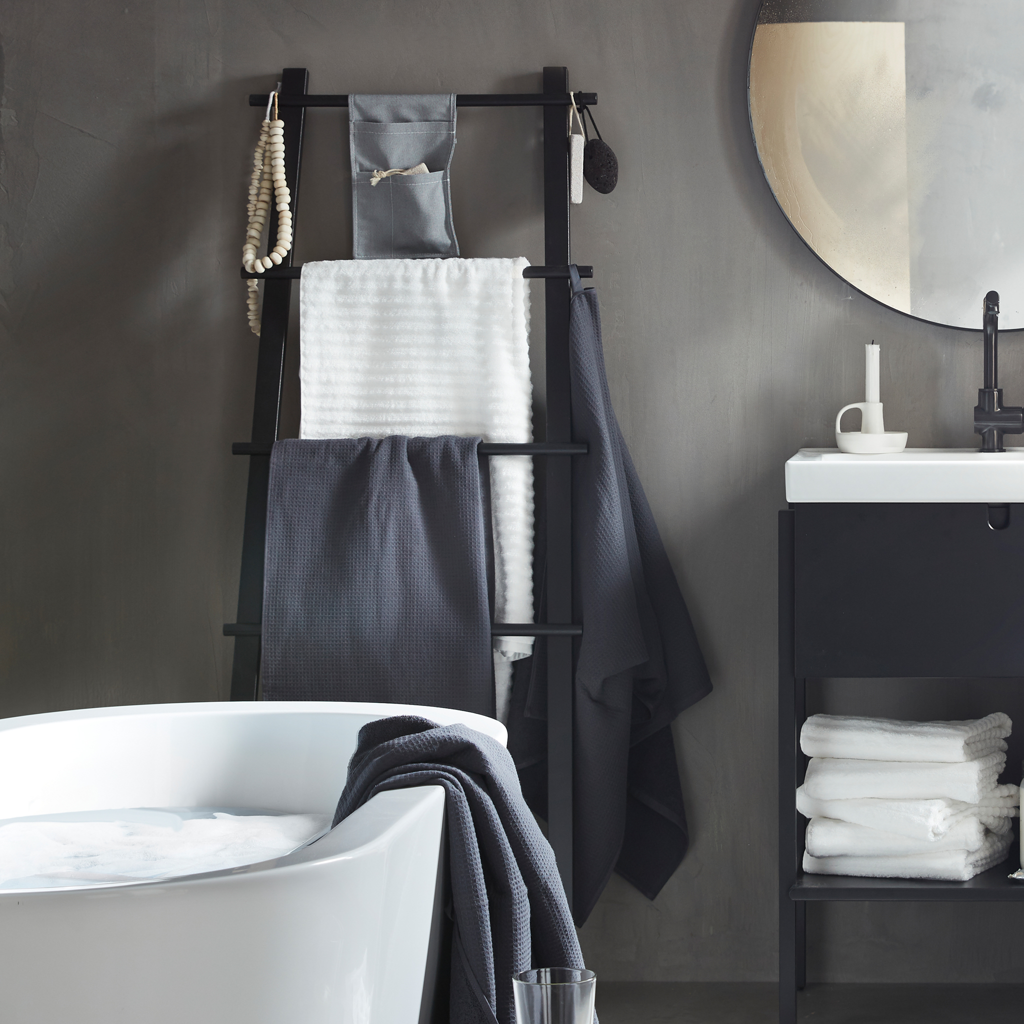 A black ladder-like VILTO towel stand in wood leaning against the grey wall of a minimalistic bathroom.