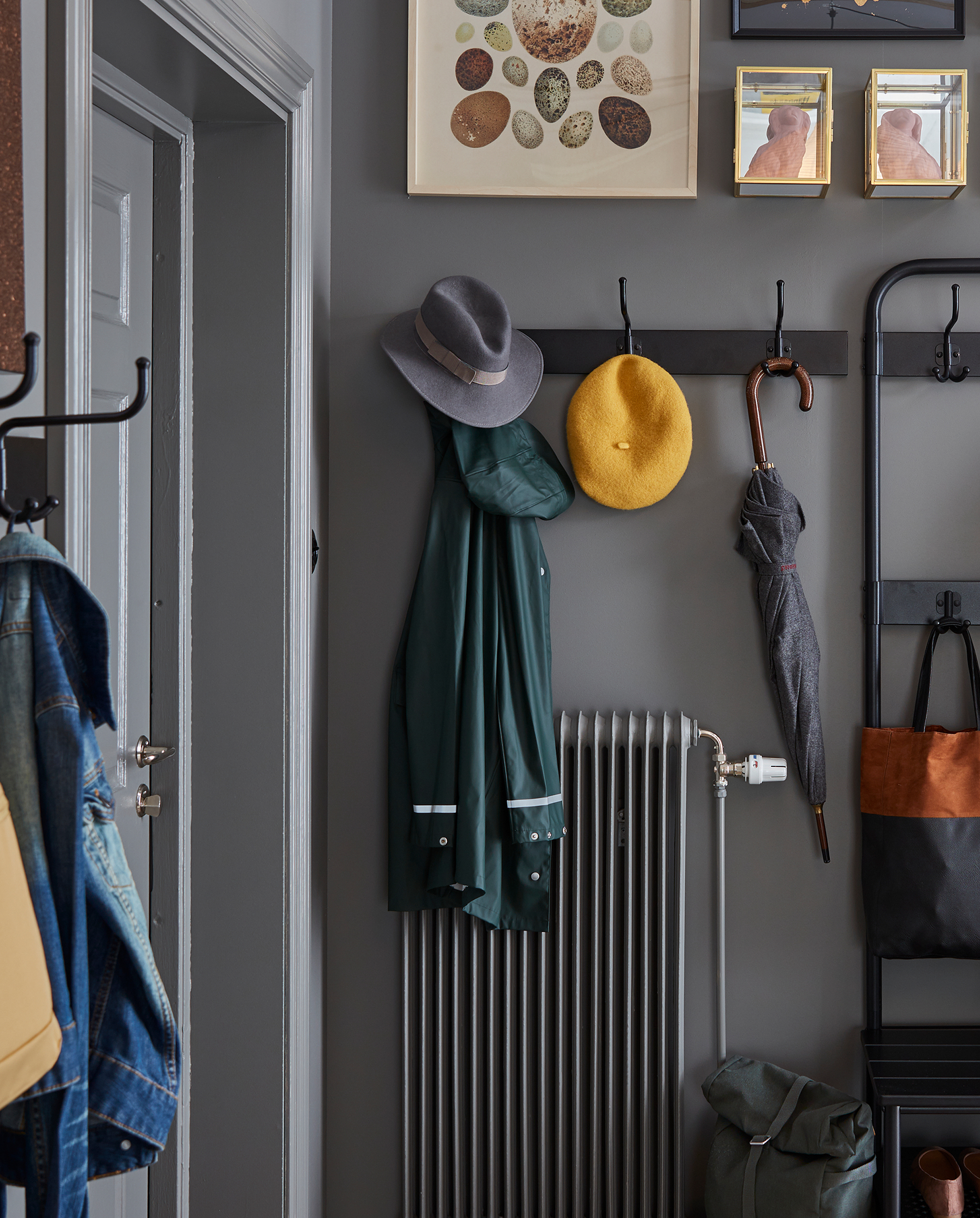 A black rack with three hooks is wall-mounted over a radiator. An umbrella, two hats and a rain jacket hang on the hooks.