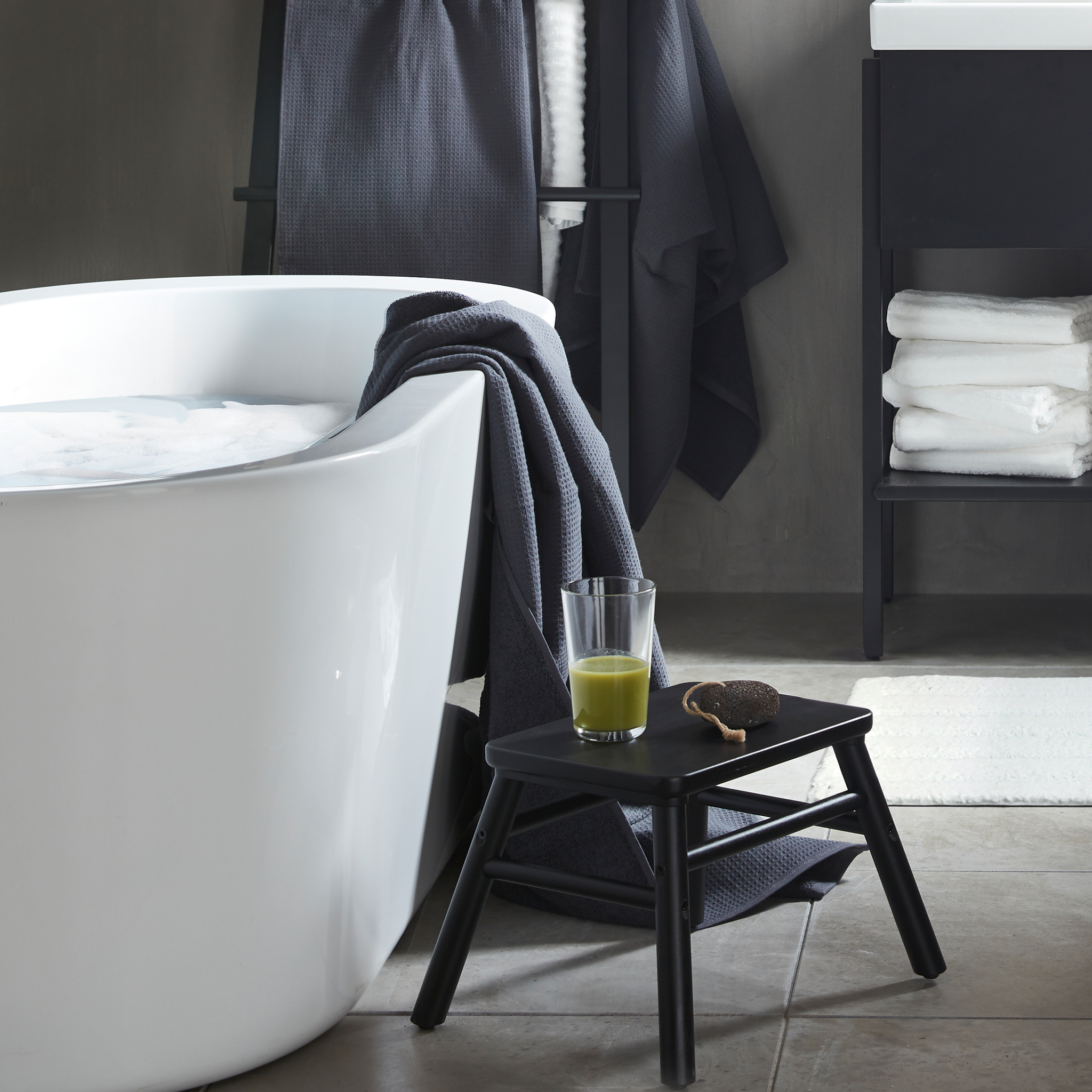 A black VILTO step stool in wood placed beside a bathtub with a glass of green smoothie on it in a minimalistic bathroom.