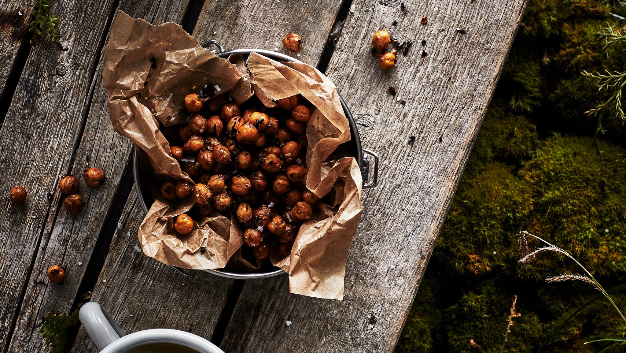 A bowl lined with paper on a rustic wooden table, filled with toasted chai and chilli chickpeas. A cup is nearby.
