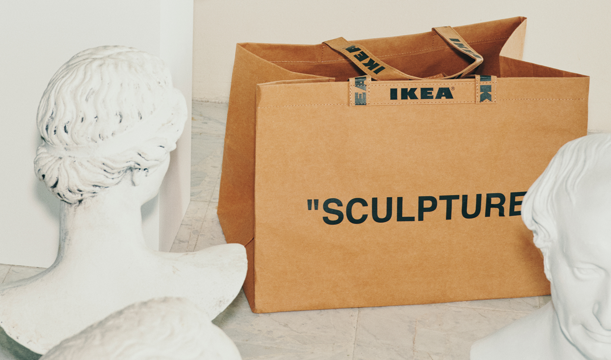 A brown IKEA bag, with the word “SCULPTURE” written on it, sits between two white sculptures.