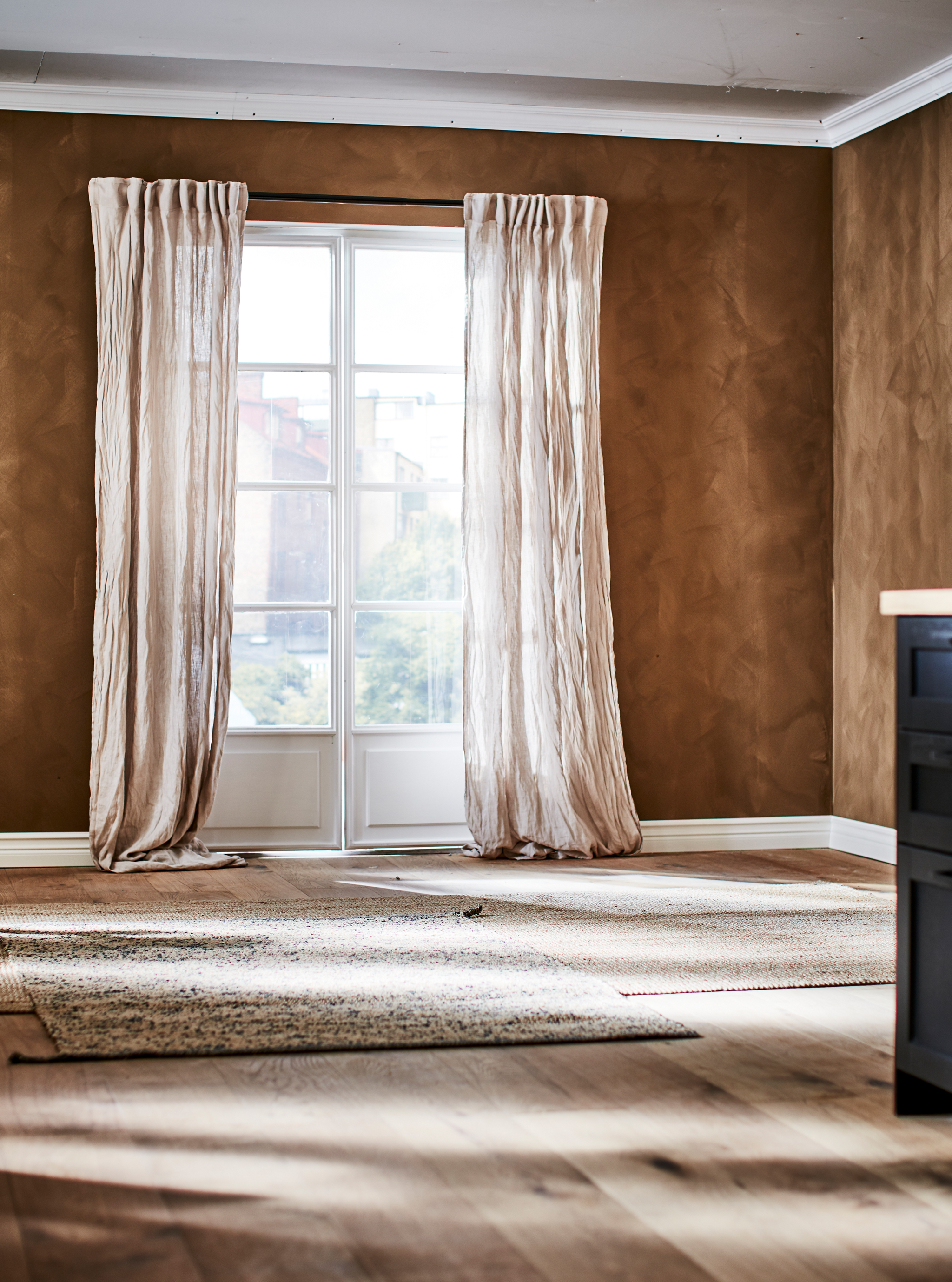 A brown room with French windows with crinkled-linen curtains, and light-brown, natural-material rugs on the wooden floor.
