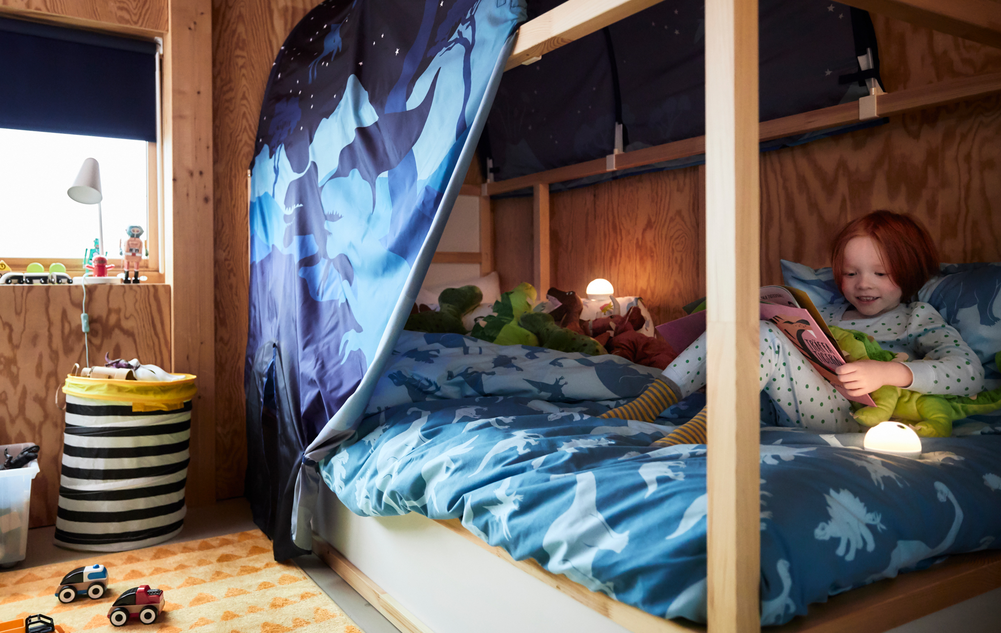 A child cuddles up with her toy dinosaur on her KURA bed, reading a storybook under a bed tent also covered in dinosaurs.