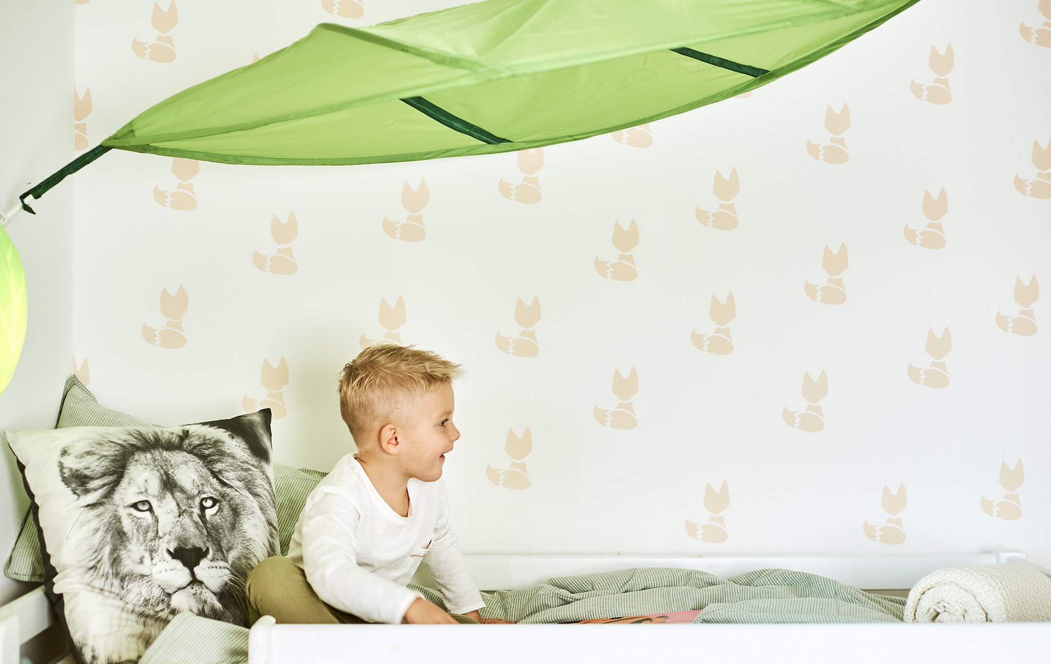 A child sits on a bed with green duvet and a cushion printed with a lion’s face, above the bed is a green leaf-shaped canopy.