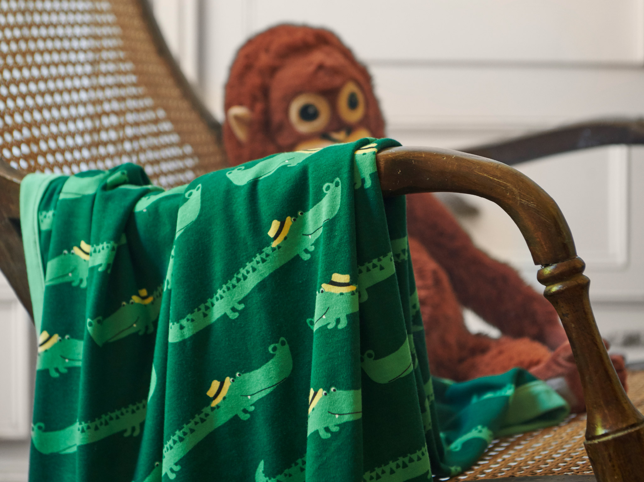 A close-up of RÖRANDE blanket with friendly crocodiles crawling across the green background, placed over a wicker chair.