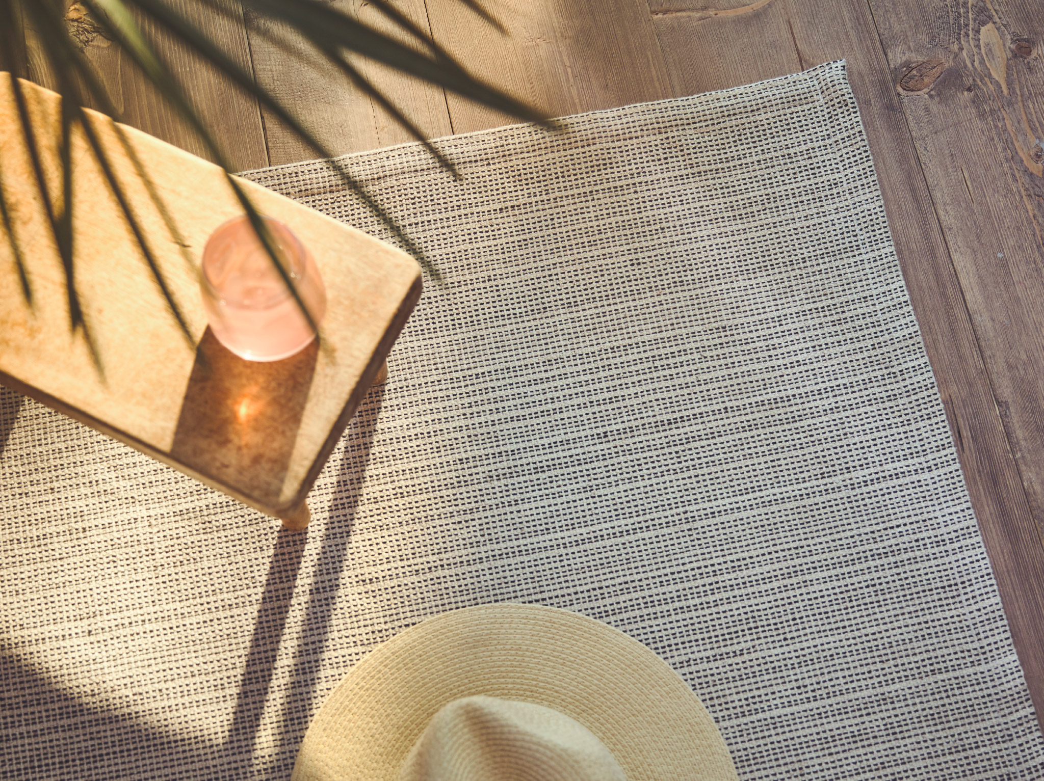 A close-up of the flatwoven TIPHEDE rug with a side table and Panama hat placed on it, while sunlight streams in on one side.