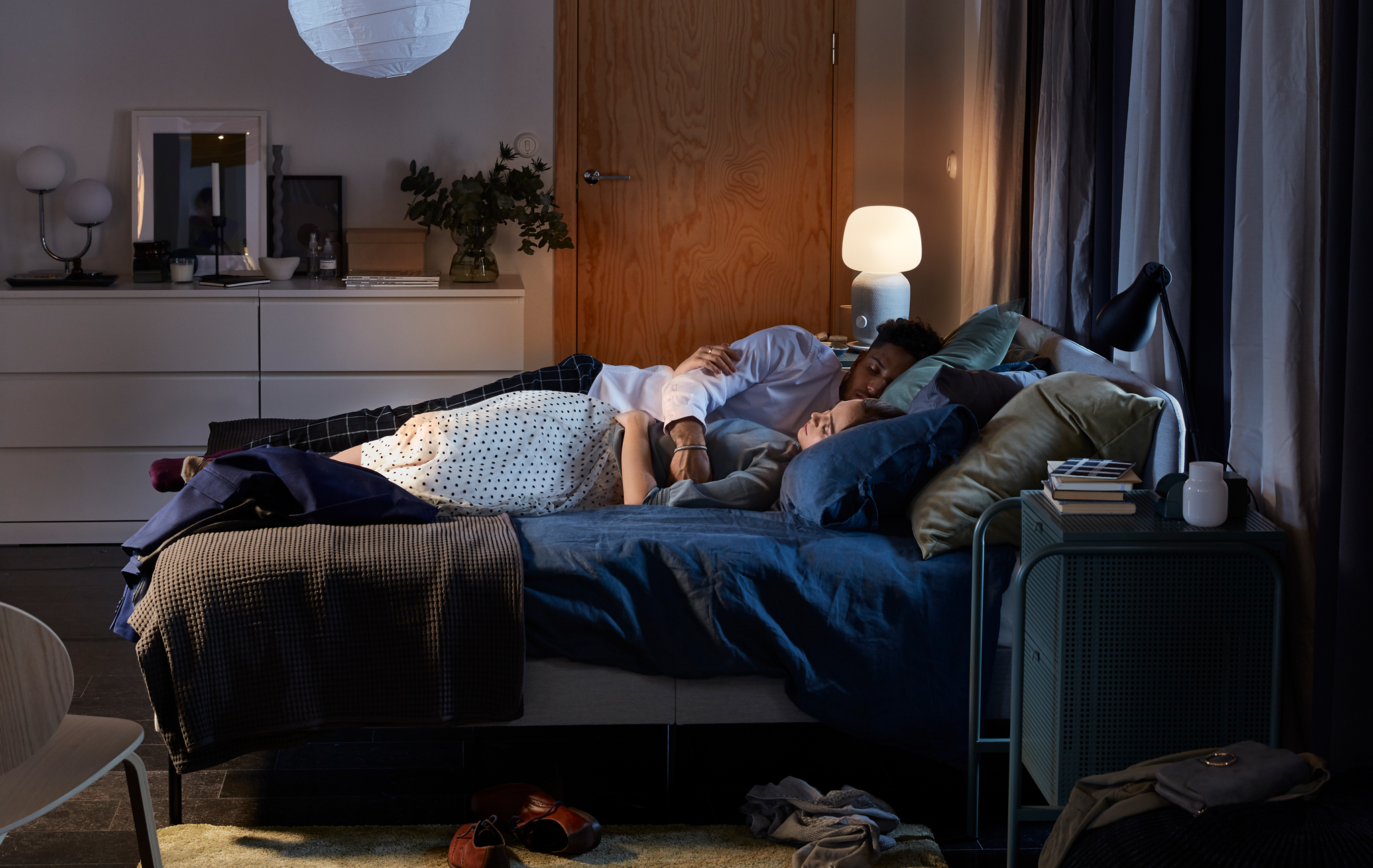A couple cuddle asleep in a SLATTUM upholstered bed, with a SYMFONISK lamp/speaker lit in the background.