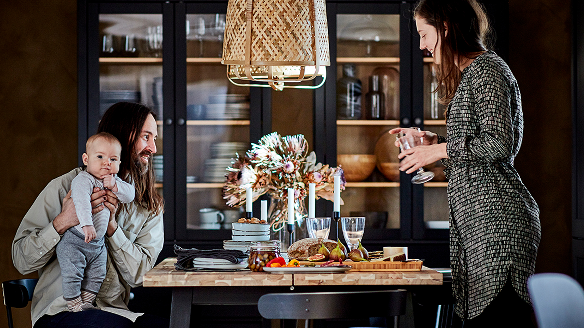 A couple with a baby gathered around a dining table. Dark glass-door cabinets with table accessories by the far wall.
