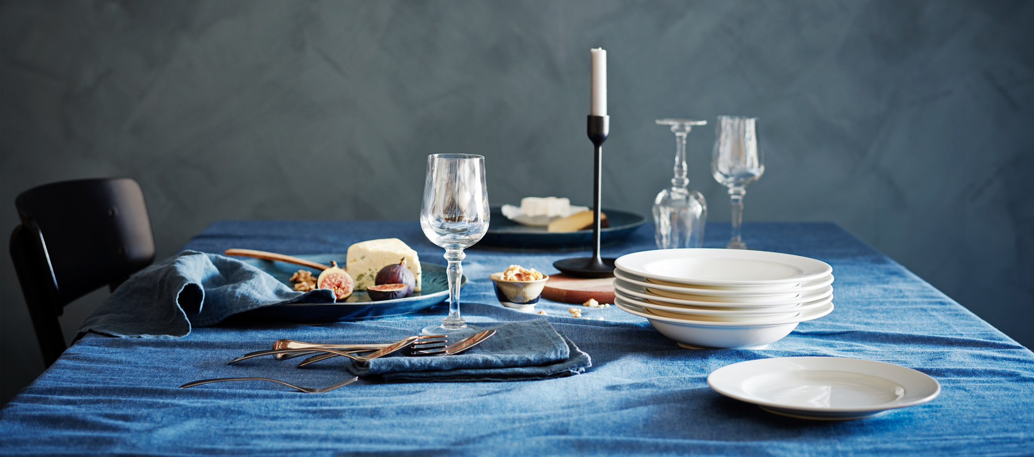 A dining, elegantly table set with KONUNGSLIG wine glass, plates, dishes, a candle stick, some food and a blue table cloth.