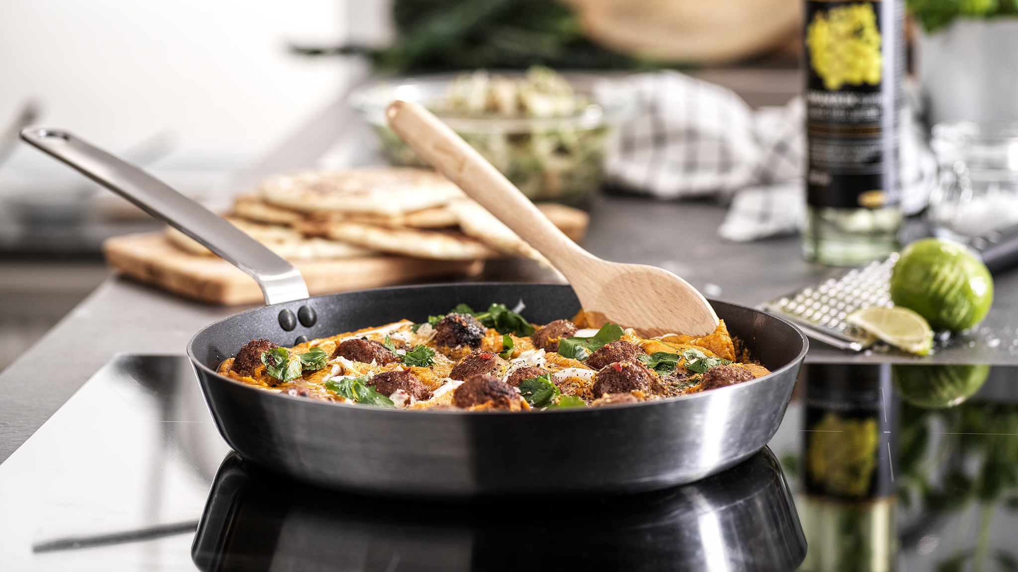 A frying pan with a handle in stainless steel is standing on a hob cooking meatballs in sauce. A wooden spoon is on the side.