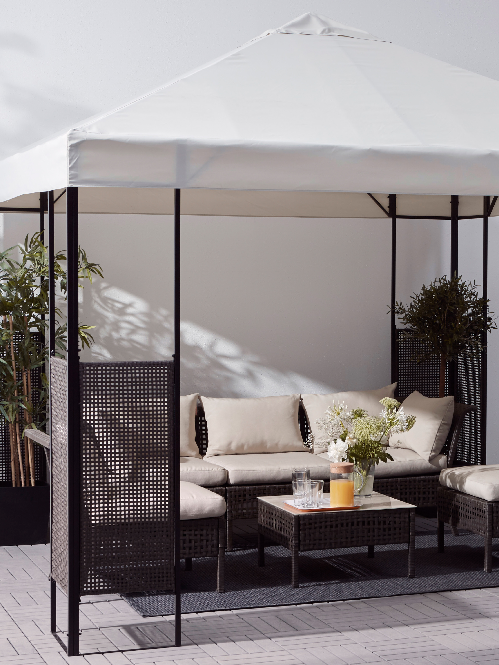 A gazebo with a white roof and dark brown frame, an outdoor sofa and chairs with cushions, and a table with plants.