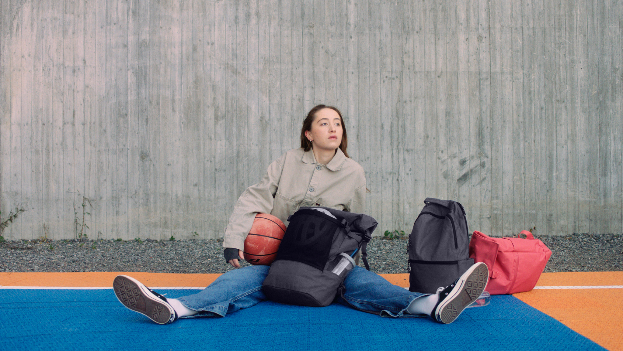 A girl holding a basketball, sitting on a basketball court with DRÖMSÄCK, VÄRLDENS and STARTTID bags lying next to her.