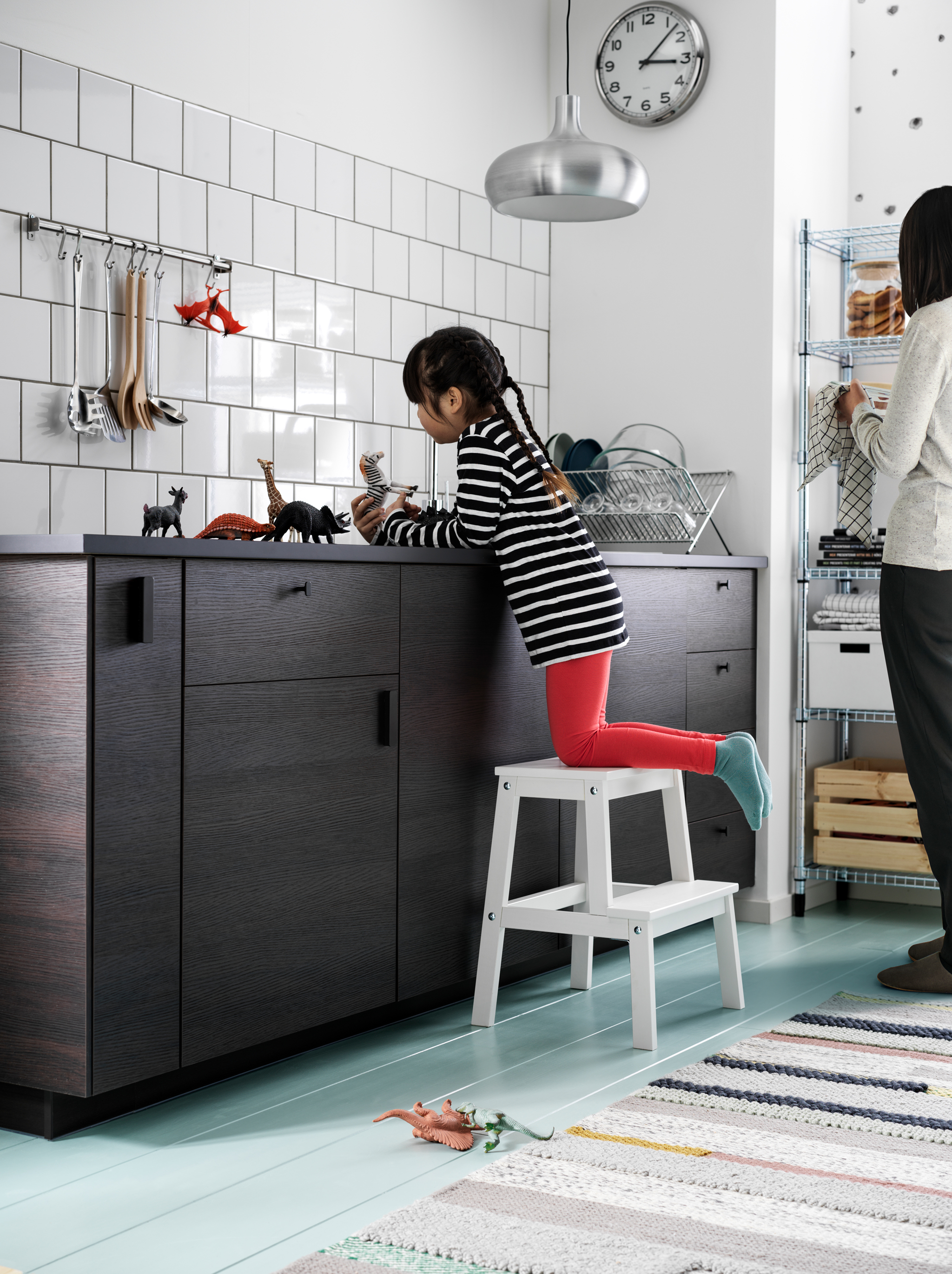A girl, perched on a BEKVÄM step stool, plays with small toy animals on a dark kitchen worktop. A woman stands nearby.