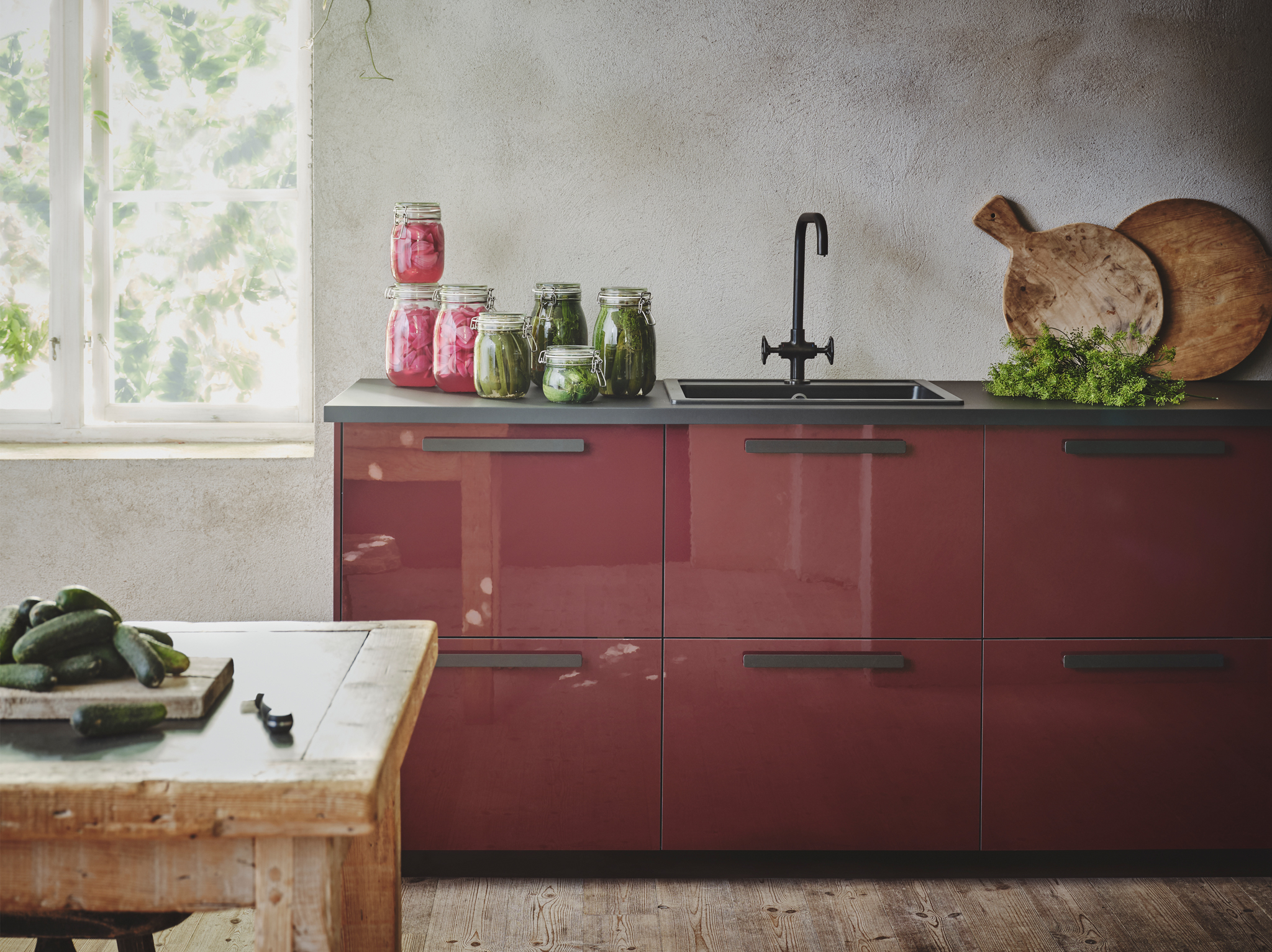 A glossy dark red-brown kitchen with black countertops, handles and sink, stands in a rustic looking room with grey textured walls.