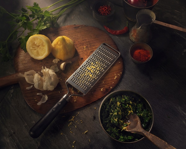 A grater in stainless steel with a black plastic handle. Shown together with a lemon on a wooden chopping board.