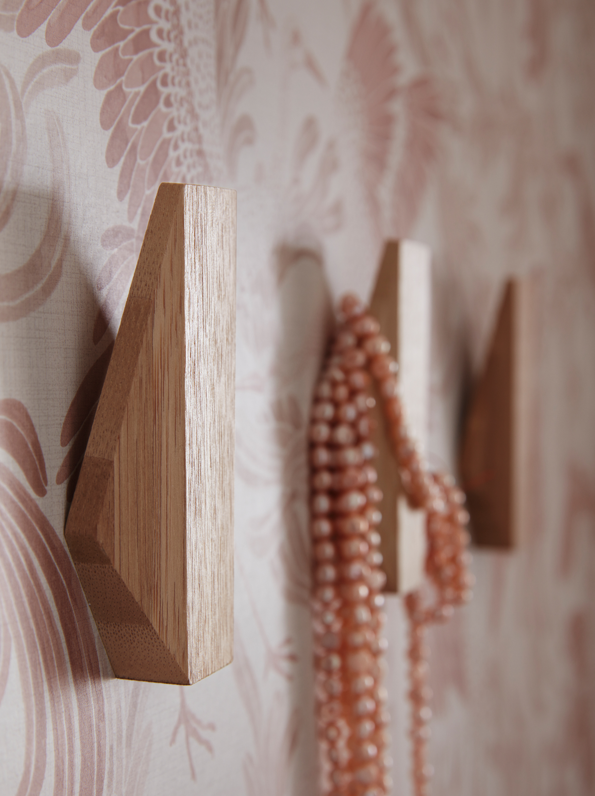 A hallway wall with three SKUGGIS geometrically shaped wooden hooks and a necklace hanging on one of them.