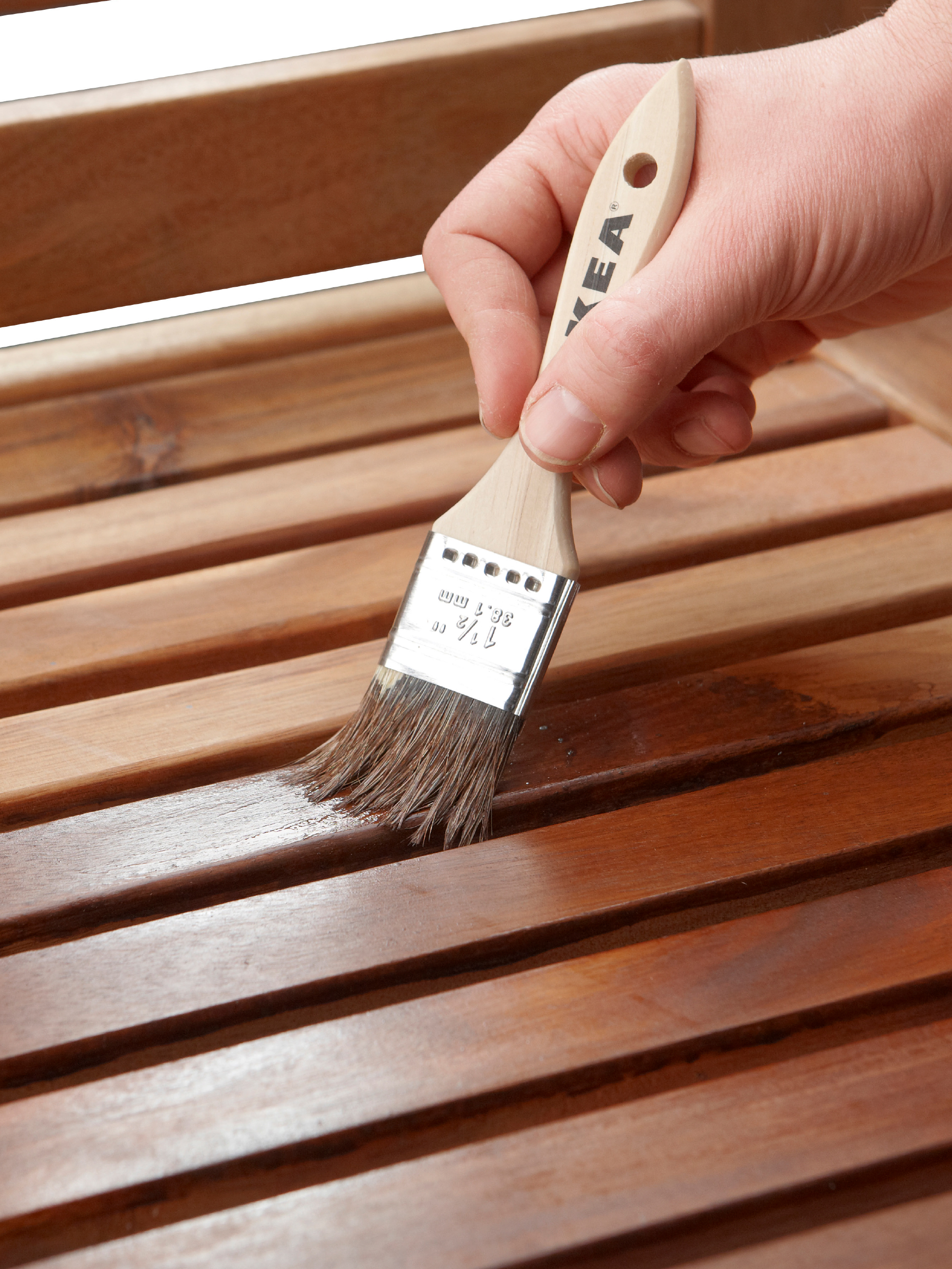 A hand holding a paint brush, painting the wooden slats on a piece of outdoor furniture with stain.