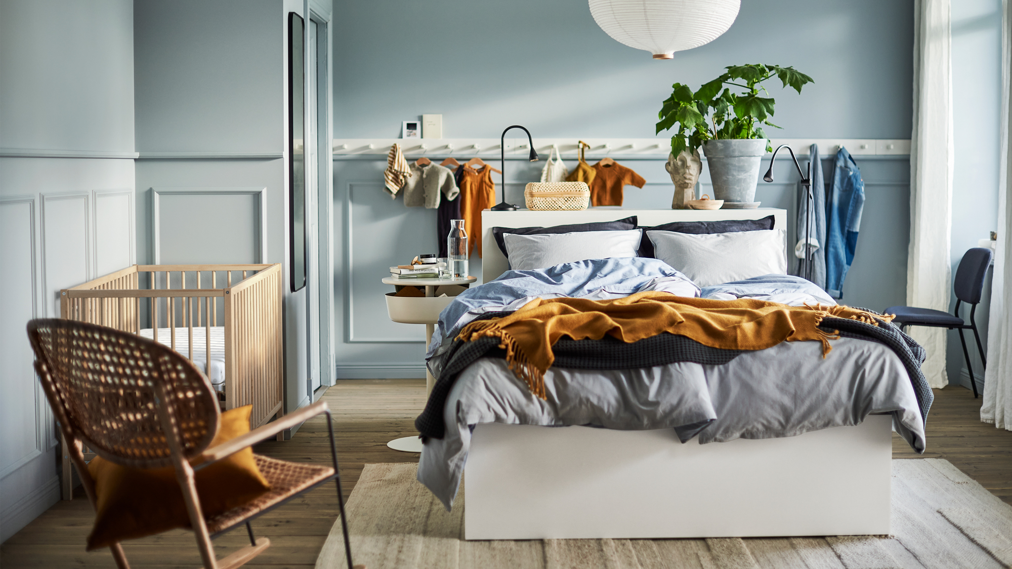 A light blue bedroom with a white bed, a white lamp, a white panel of hooks, a rattan rocking chair and a SNIGLAR baby cot.
