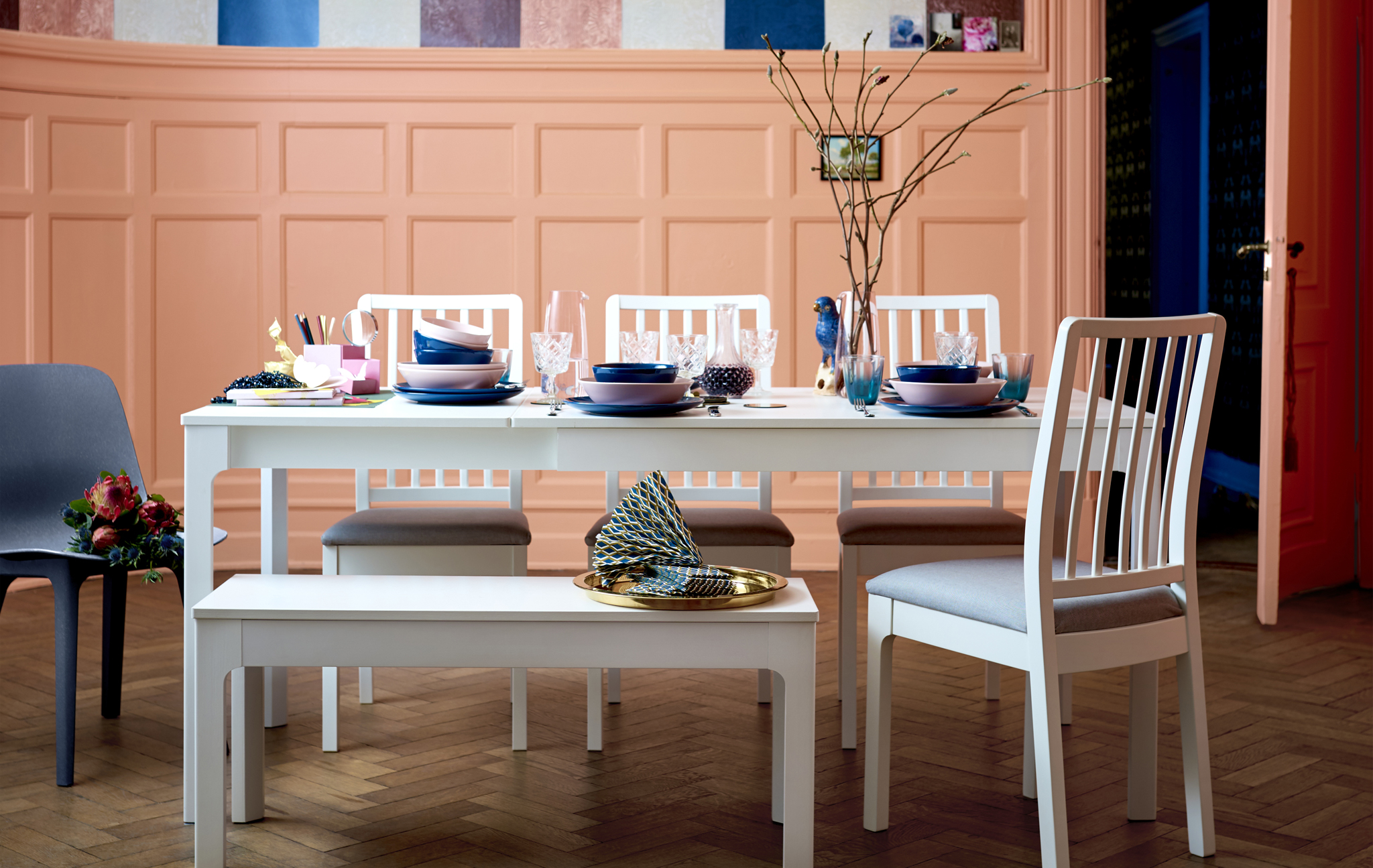 A long white dining table, chairs and bench set with colourful tableware.