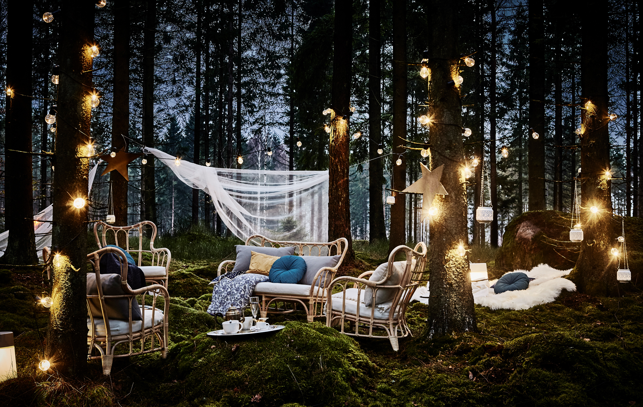 A lounge area at night in a forest wedding with rattan armchairs, lots of cushions, lighting chain and hanging lanterns