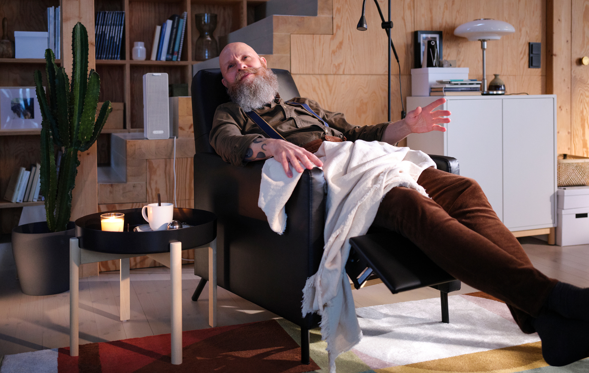 A man leans back under a blanket in a black GISTAD recliner, listening to music on a white SYMFONISK WiFi speaker.