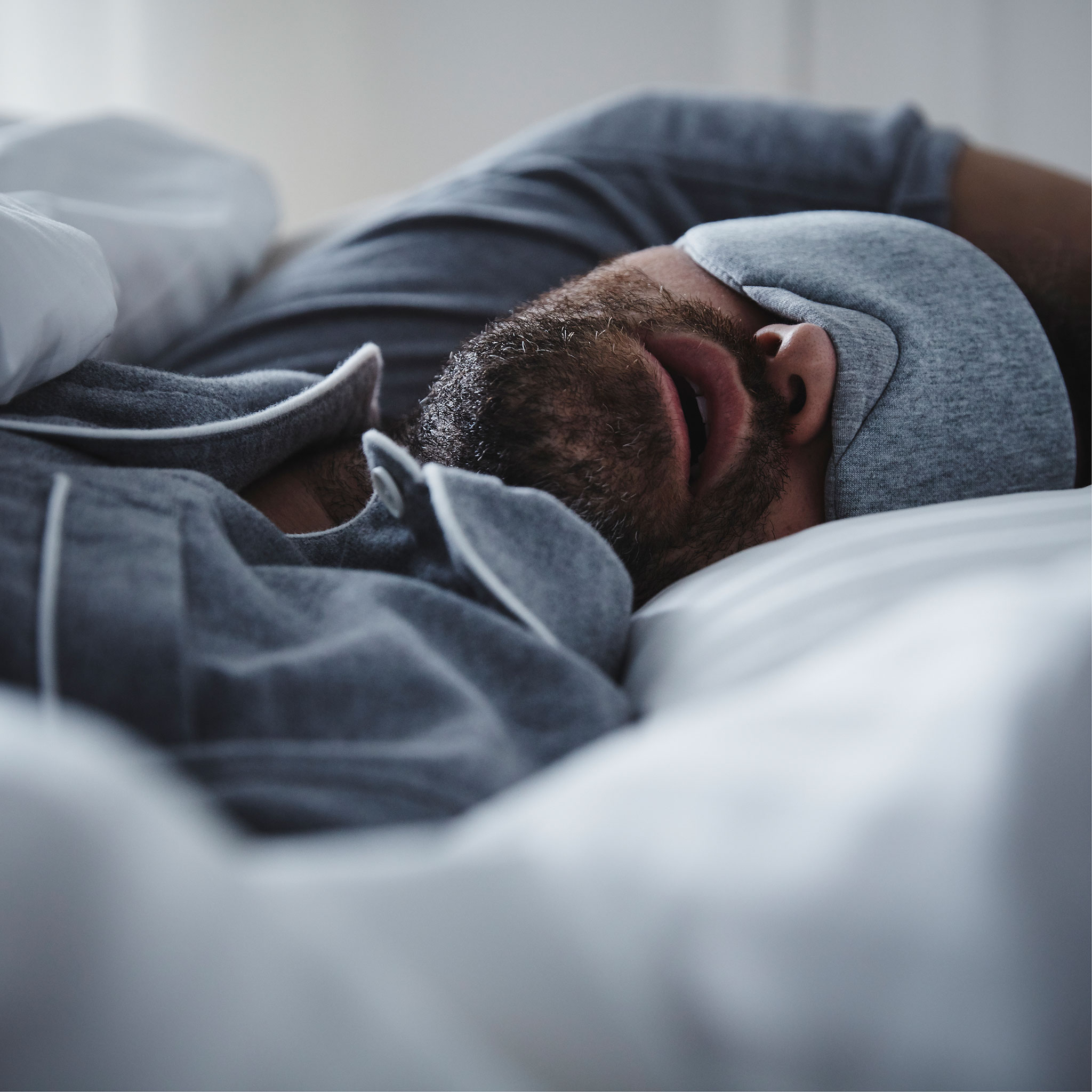 A man wearing blue pyjamas and matching eye-mask lies asleep in bed with his mouth open.