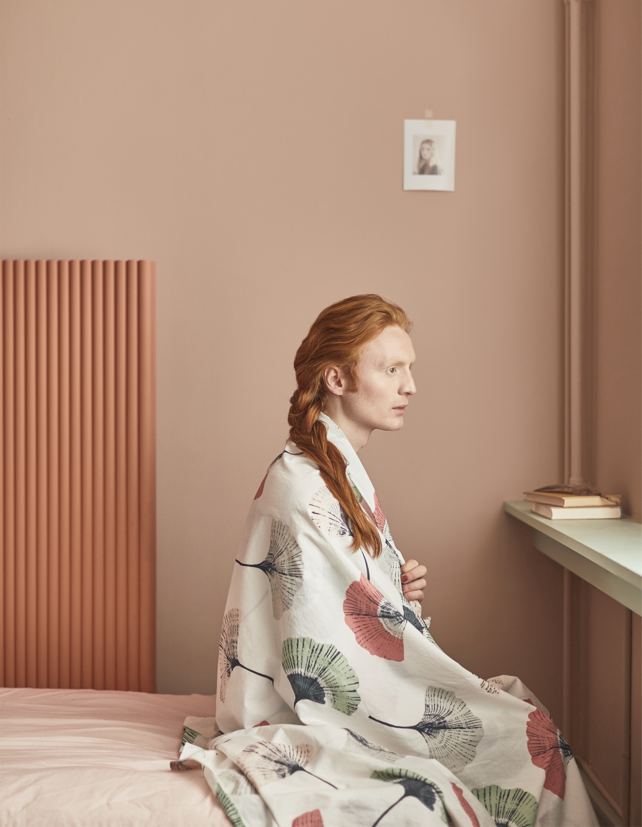 A man with red hair sitting on a bed with fabric around his shoulders.