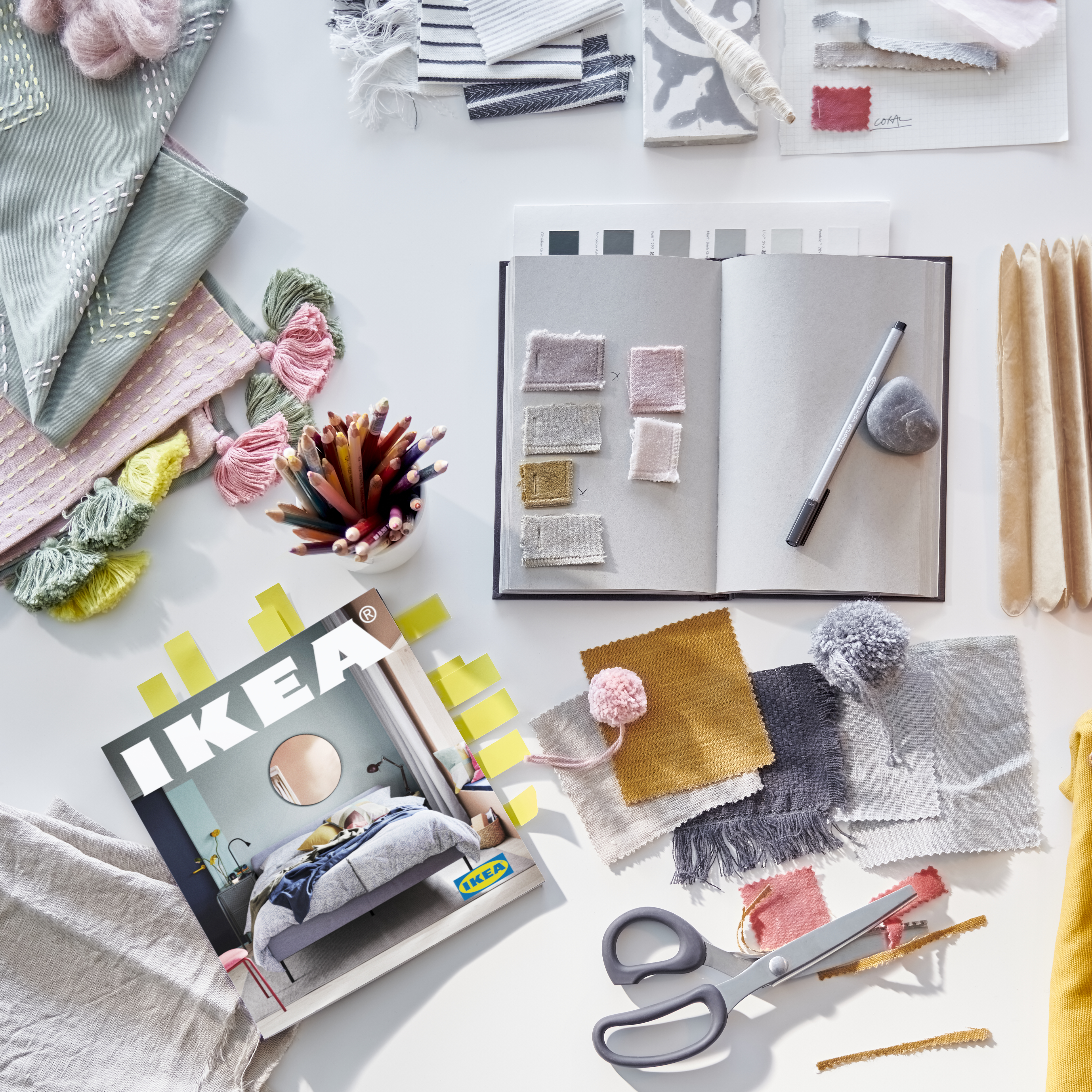 A note book, a pen, scissors, and pieces of fabric in pink, grey, yellow, mustard, and green beside a 2021 IKEA Catalogue.
