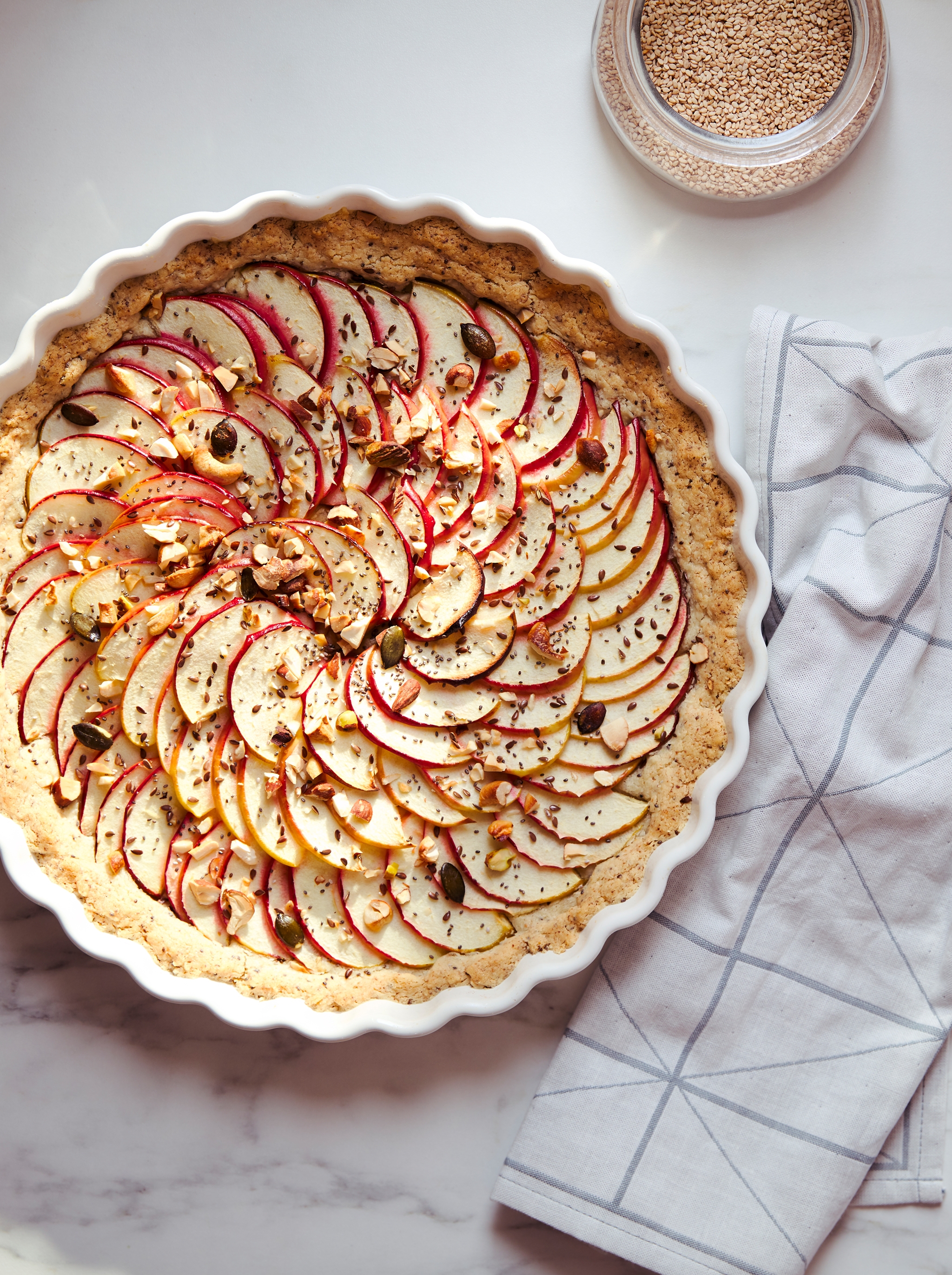 A pie covered with thin apple slices, seeds and nuts in a white VARDAGEN pie dish standing on a marble kitchen worktop.