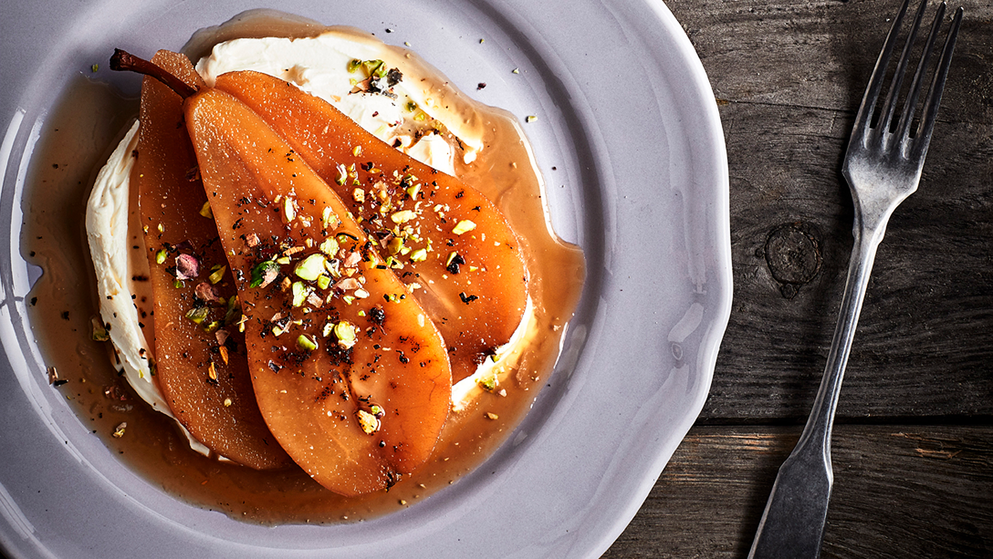 A portion of pears poached in Earl Grey tea served on a white plate on a rustic wooden table, with a fork on one side.