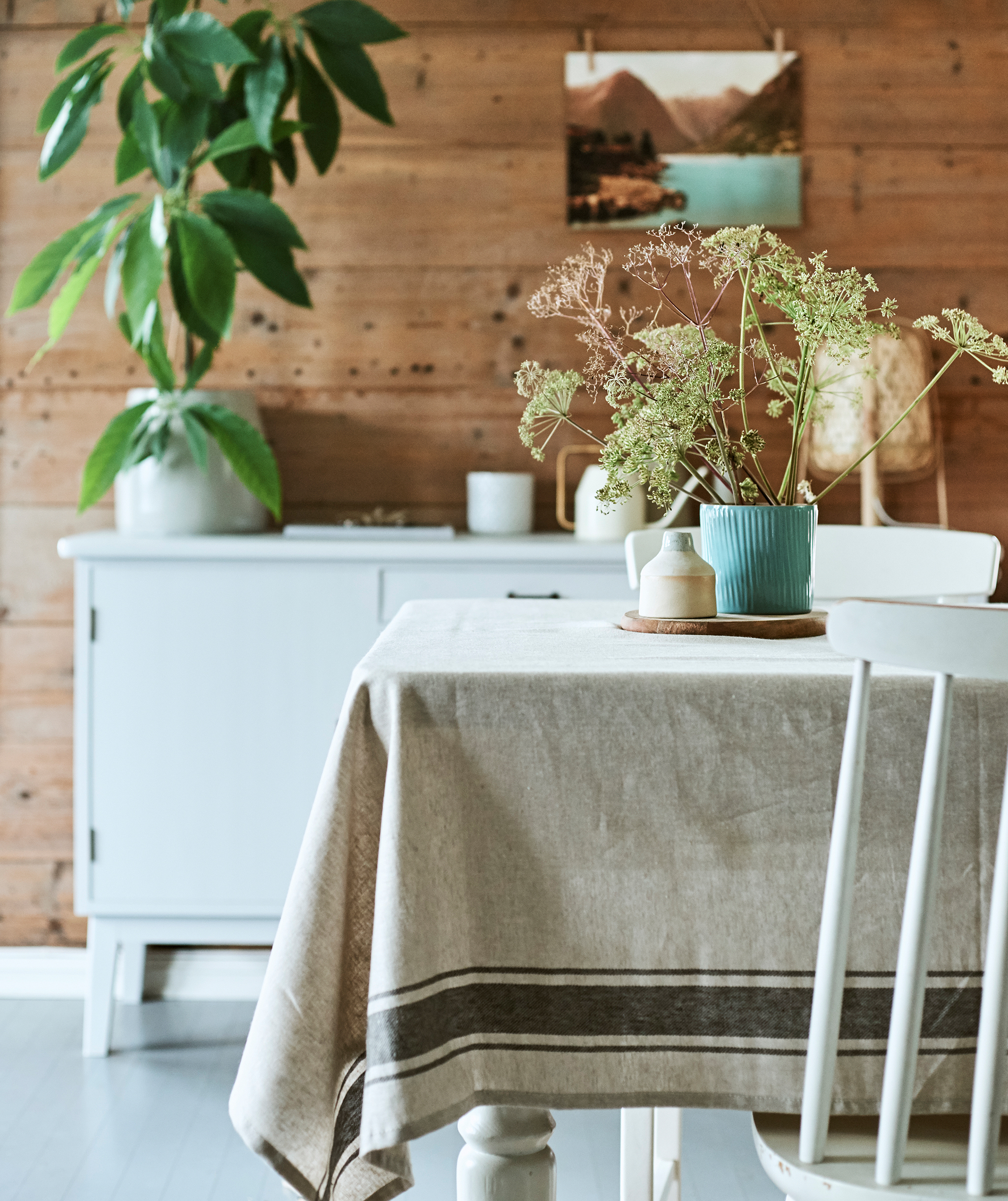 A rustic dining room with wood table covered with linen cloth and a centrepiece of wild flowers in a blue ridged plant pot.