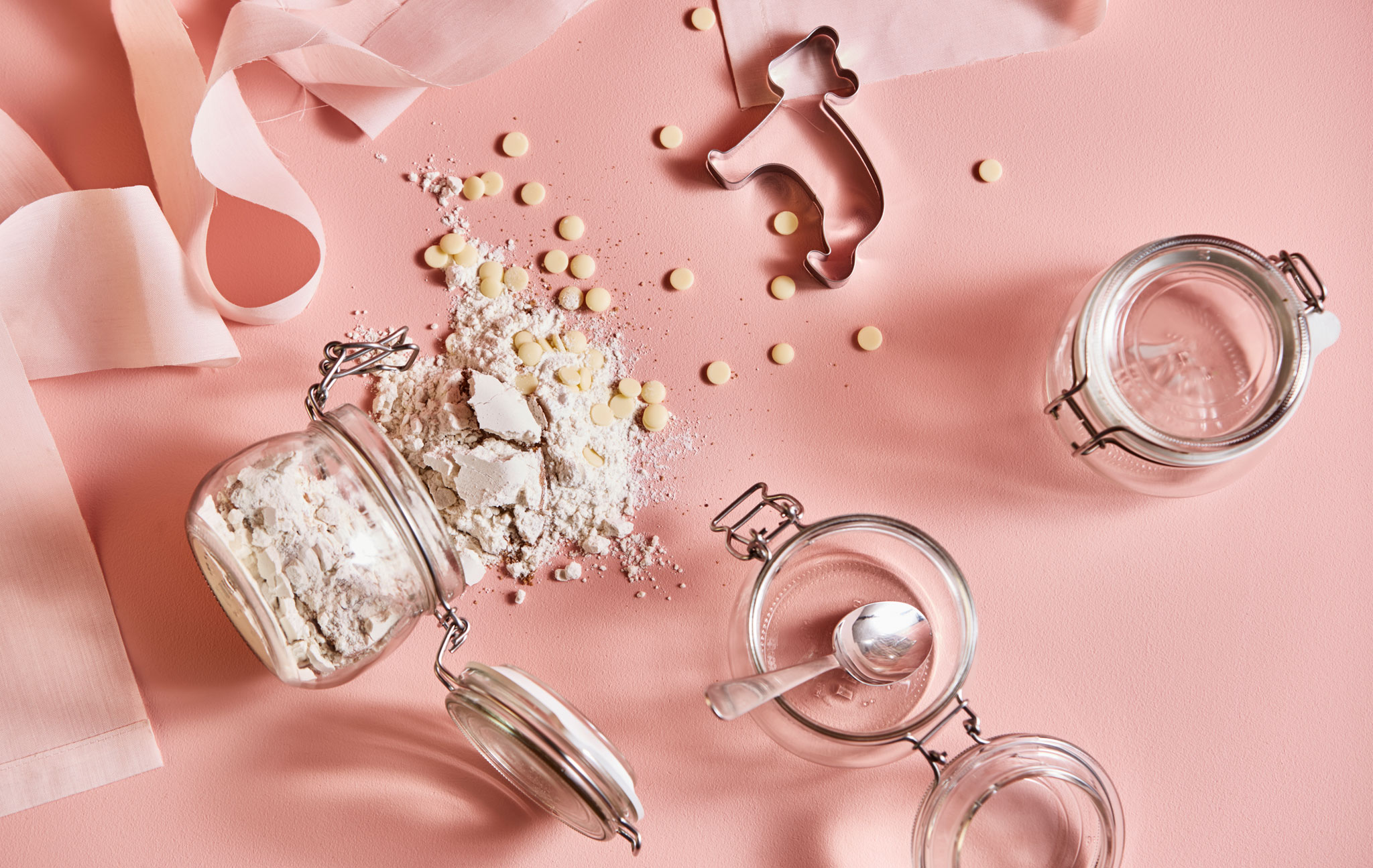 A shot from above of cookie dough ingredients, jars, and cookie cutters on a pink background.