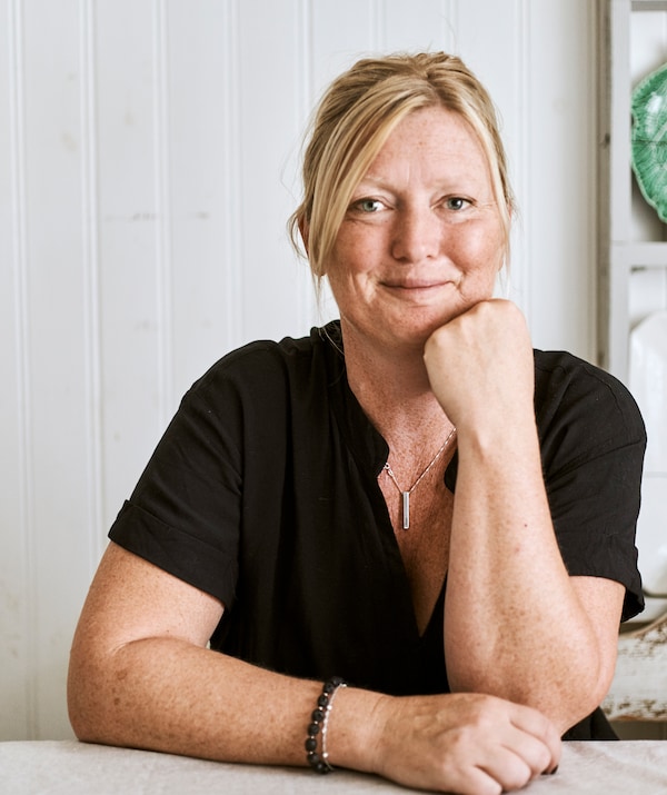 A smiling woman with blonde hair in a black shirt sits at a dining table in a room with white-washed wood panelled walls.