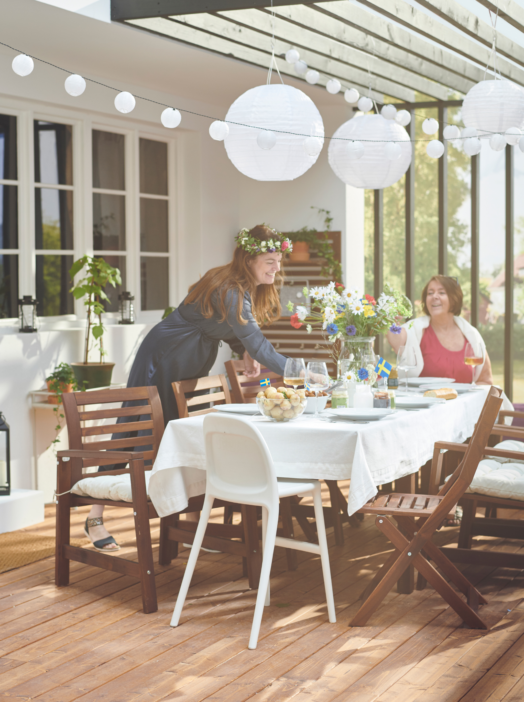 A sunlit roofed terrace with two people around a group of ÄPPLARÖ table and chairs, the table set and decorated for a party.