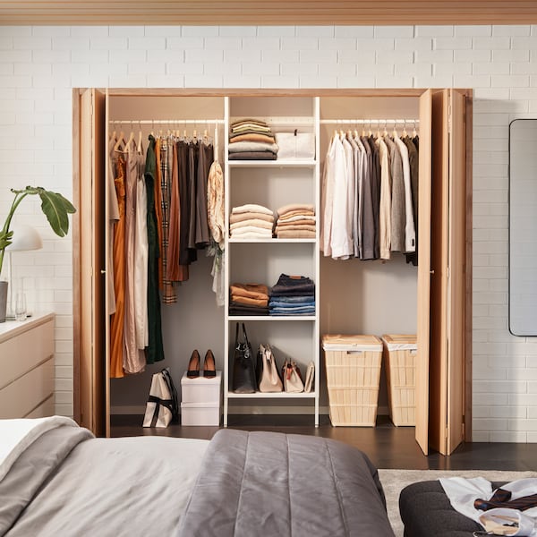 A white AURDAL wardrobe combination with shelves and rails is mounted in a space behind foldable wooden doors.
