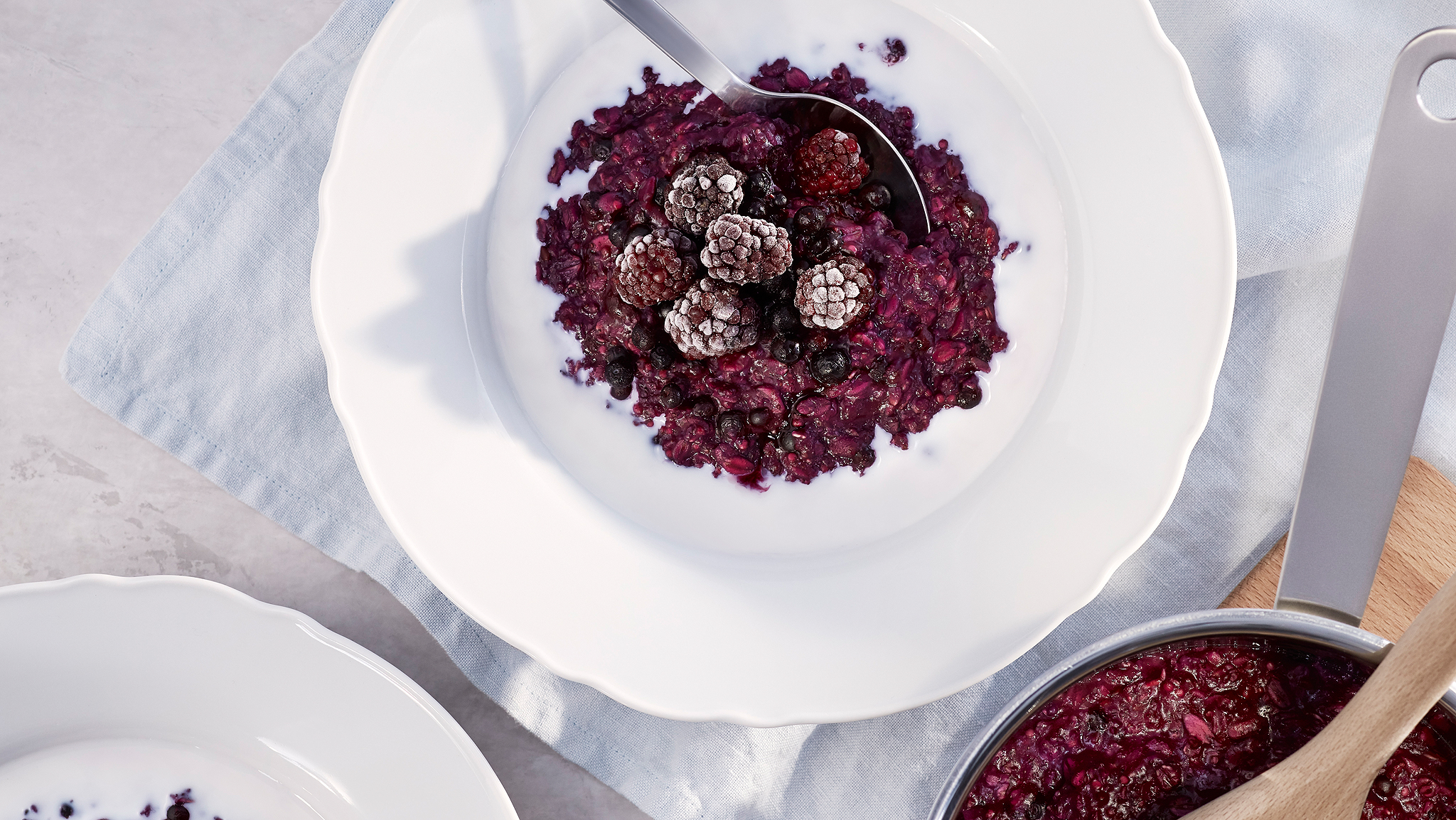A white china bowl of HJÄLTEROLL porridge and berries with a spoon in it, on top of a white napkin, beside a metal saucepan.
