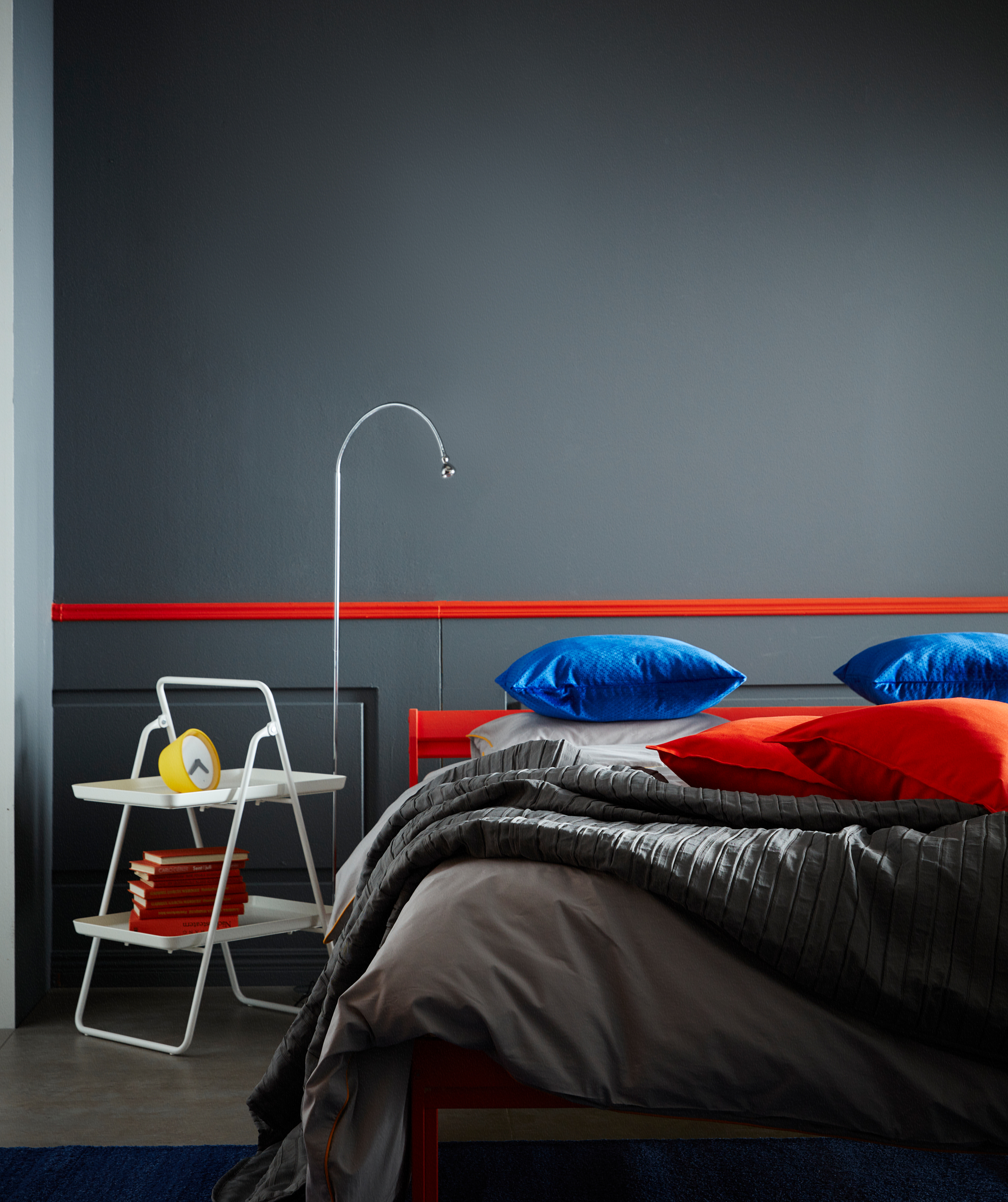 A white IKEA VIGGJA tray stand with two shelves used as a bedside table with a lamp beside it.