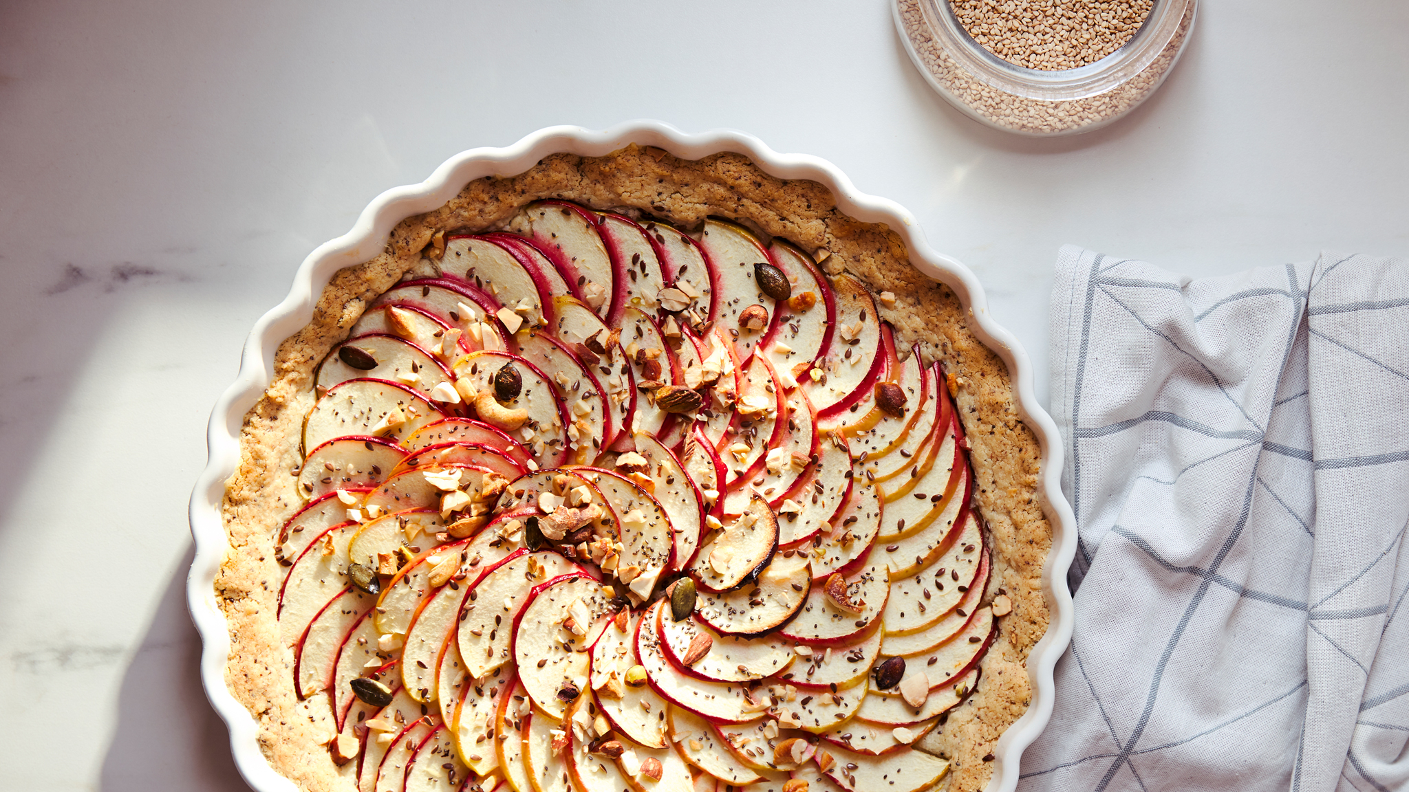 A white kitchen towel with a grey, graphic pattern and a pie covered with thin apple slices in a white VARDAGEN pie dish.